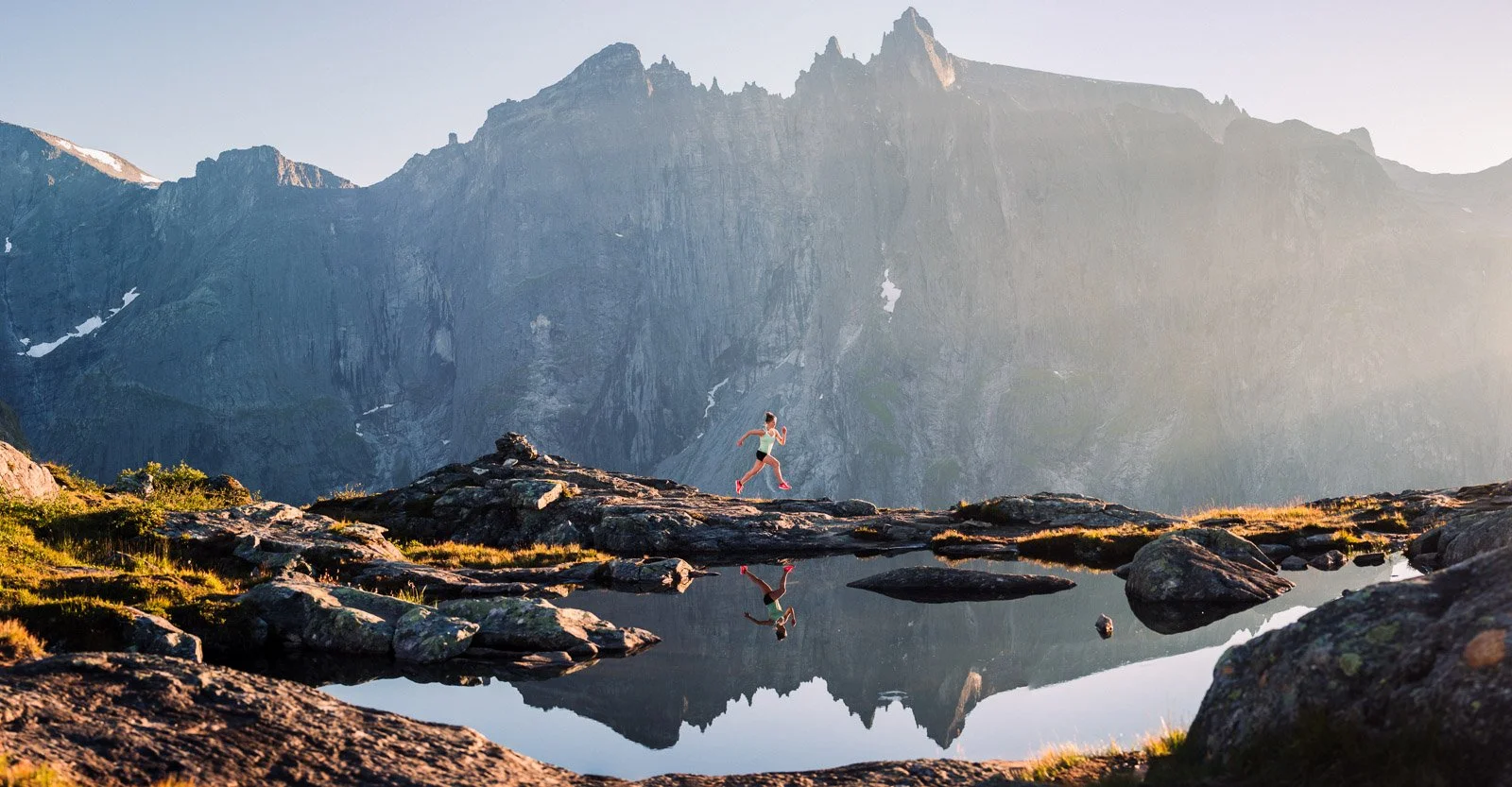 A woman running on rocky terrain near a mountain lake, with snow-capped mountains in the background and her reflection visible in the water.