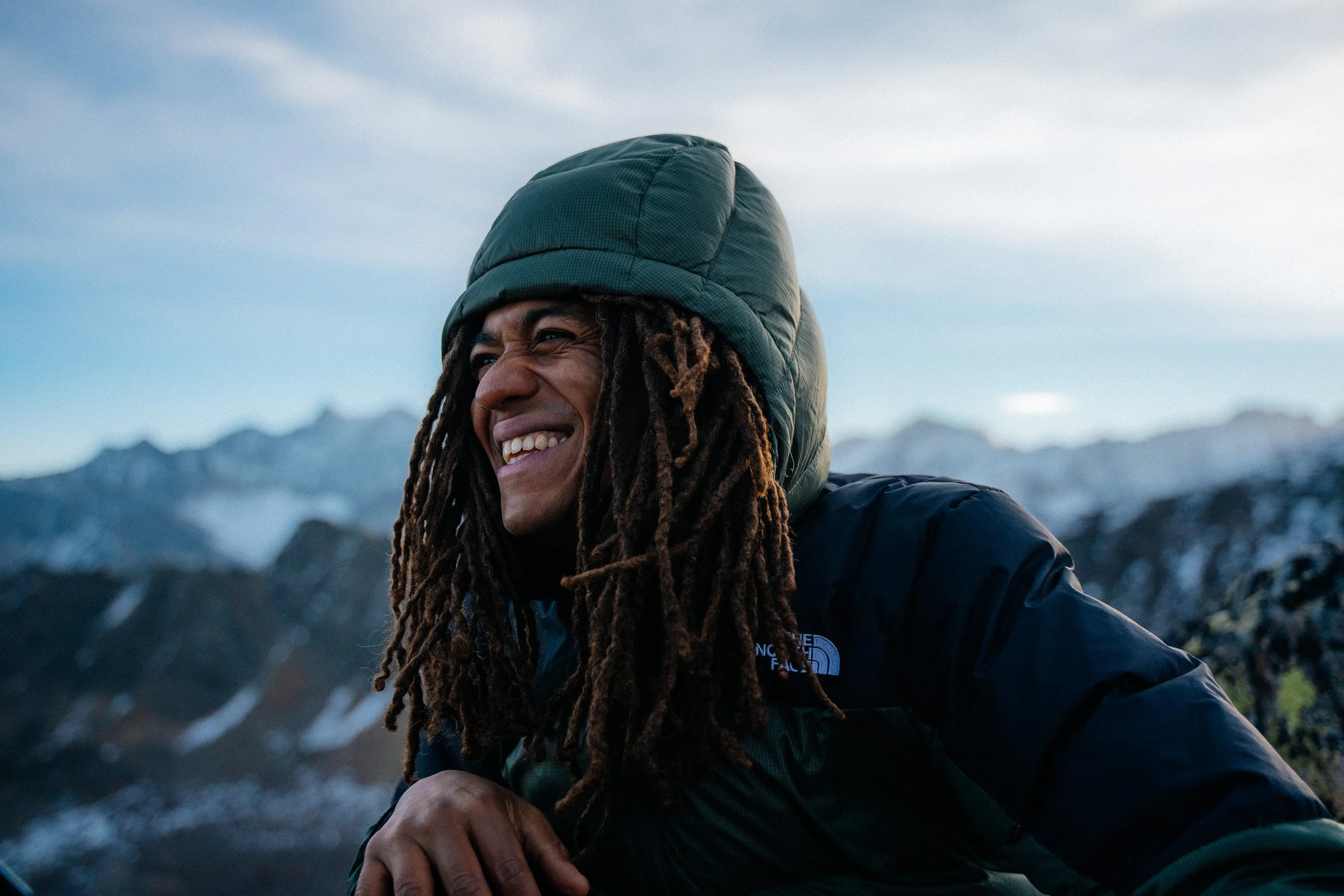 A smiling person with dreadlocks wearing a green hooded jacket outdoors with mountains in the background.