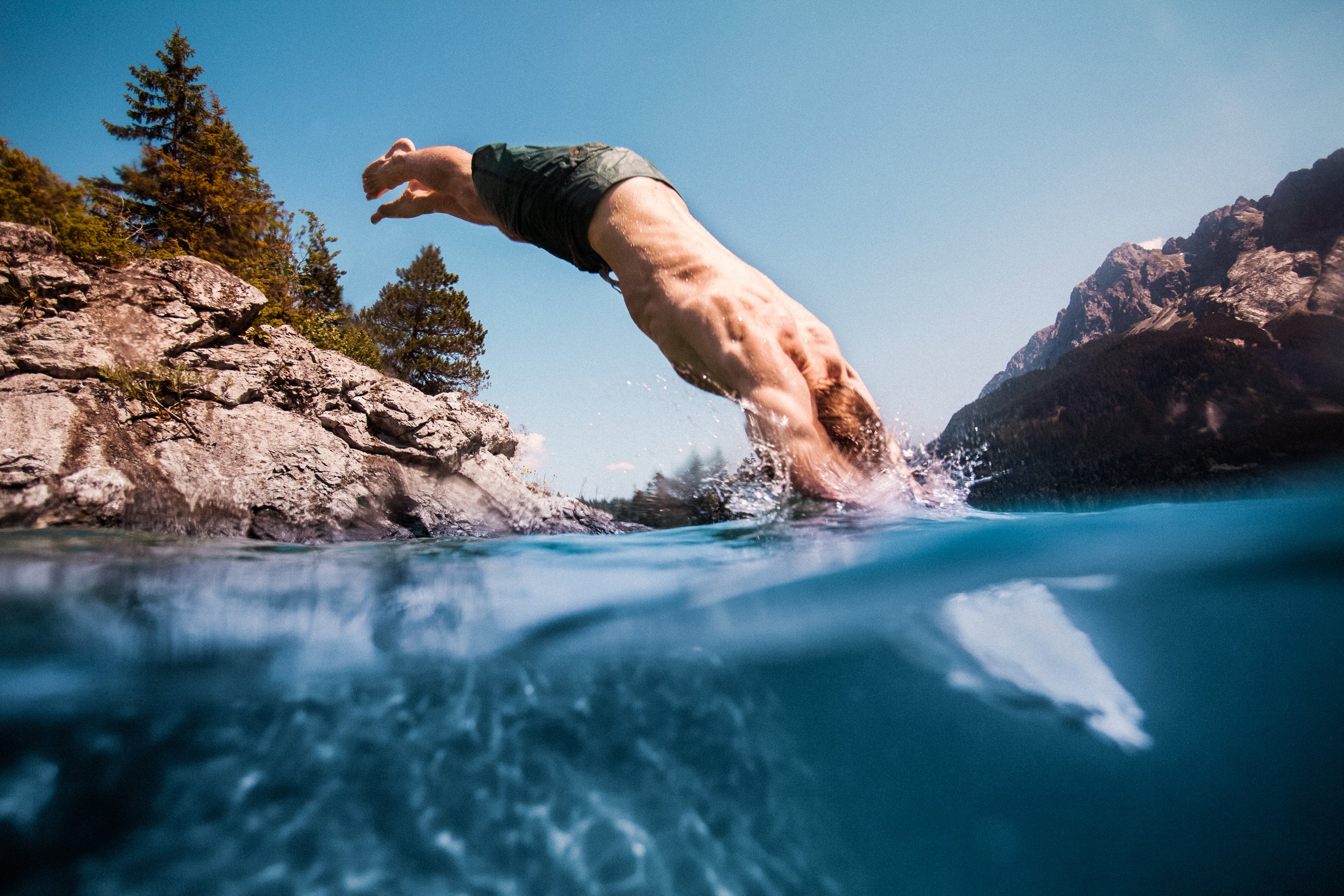 A man diving into a mountain lake from a rocky shoreline with a clear blue sky in the background.