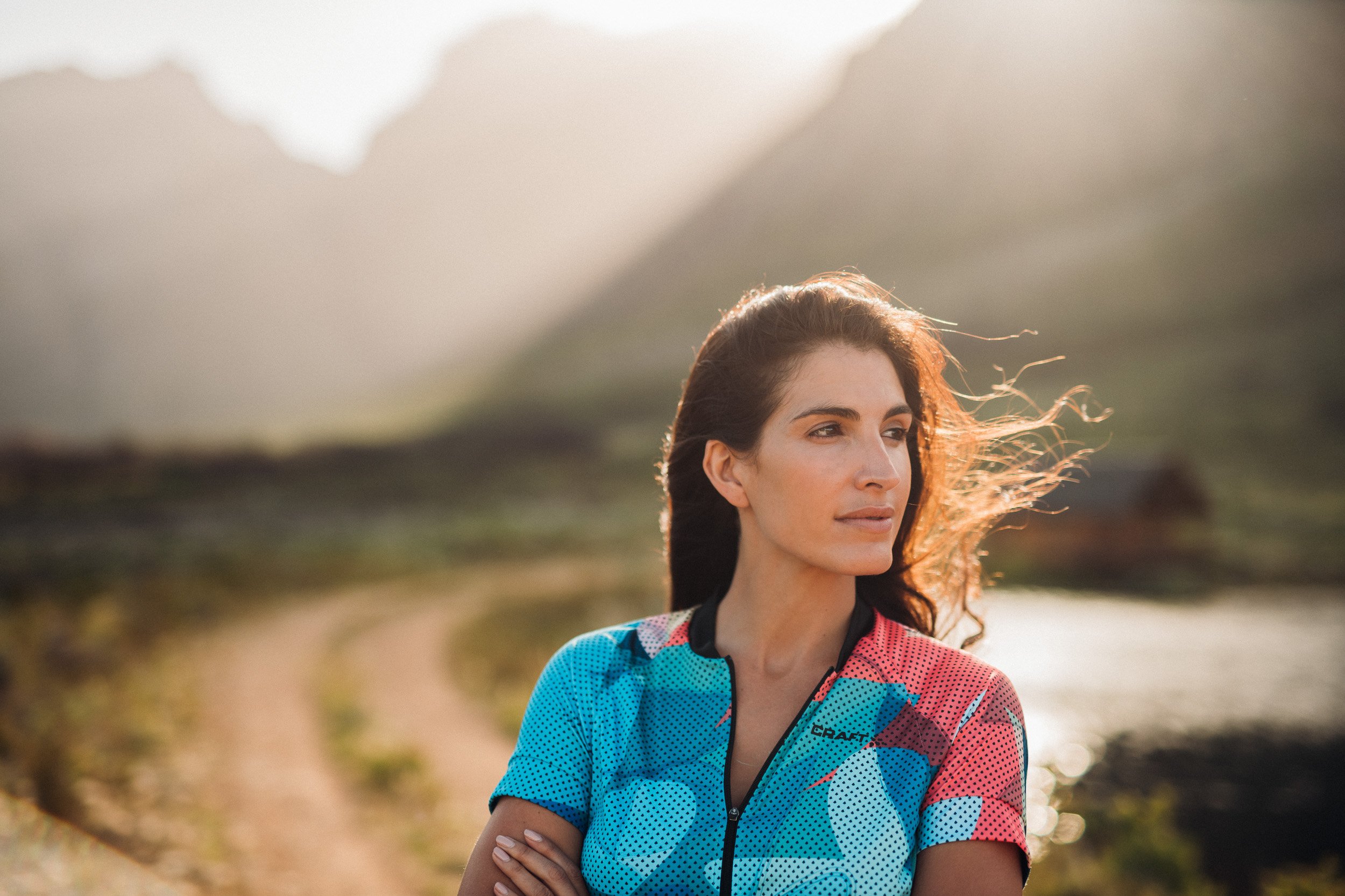 A woman with long, wavy brown hair and light skin stands outdoors near a winding dirt path and a body of water, looking to the right with a contemplative expression. She is wearing a colorful cycling jersey, and the scene is illuminated by soft sunli
