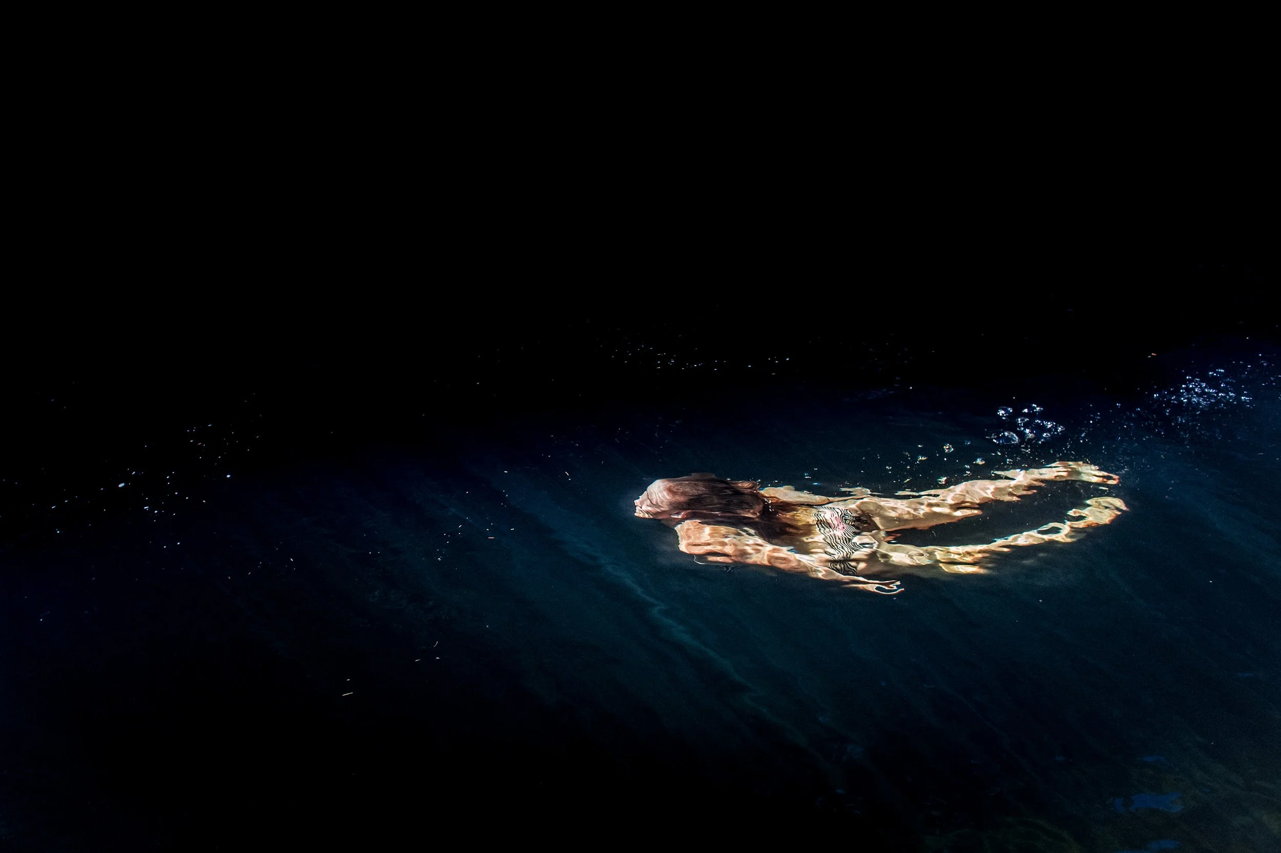 A woman floating on her back in a dark body of water, illuminated by a small light source.