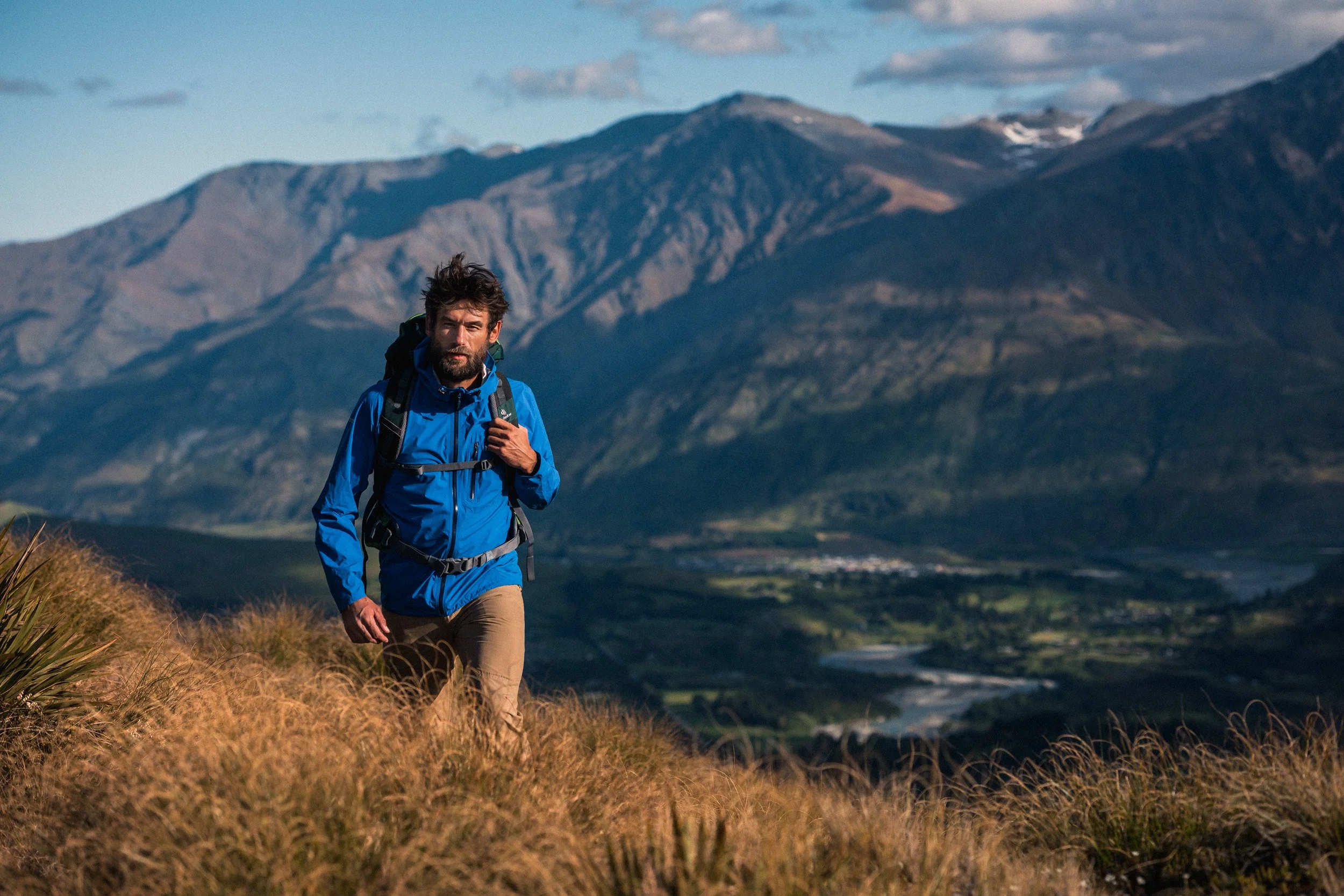 A man hiking on a mountain trail surrounded by tall grass with mountains in the background.