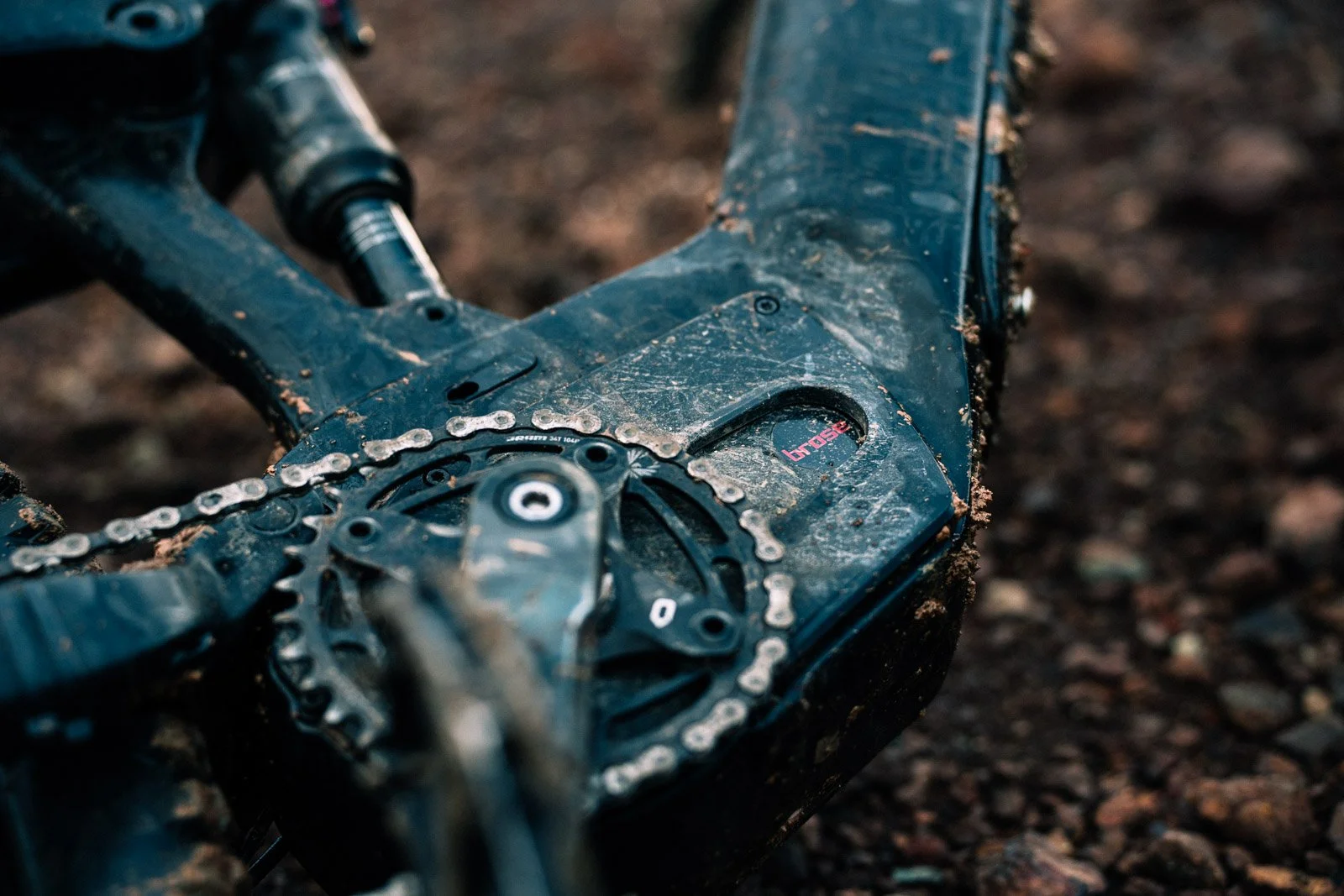 Close-up of a muddy mountain bike frame, chain, and pedal components on rough terrain.