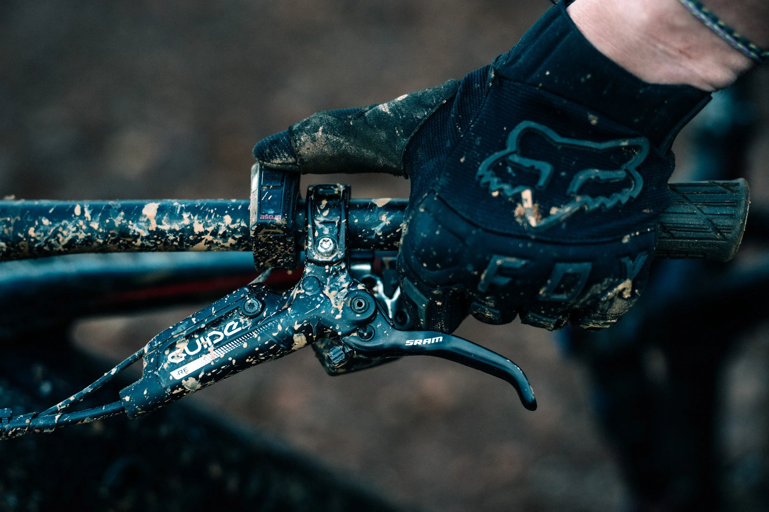 Close-up of a muddy mountain bike handlebar with a black SRAM brake lever, a handlebar with mud splatters, and a gloved hand gripping the handlebar; the glove has a Fox Racing logo.
