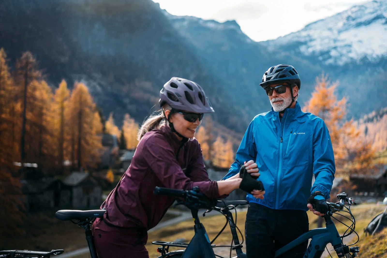 A woman and a man with bicycles in a mountainous, forested area with fall foliage. The woman is smiling and shaking hands with the man, both wearing helmets and cycling gear.