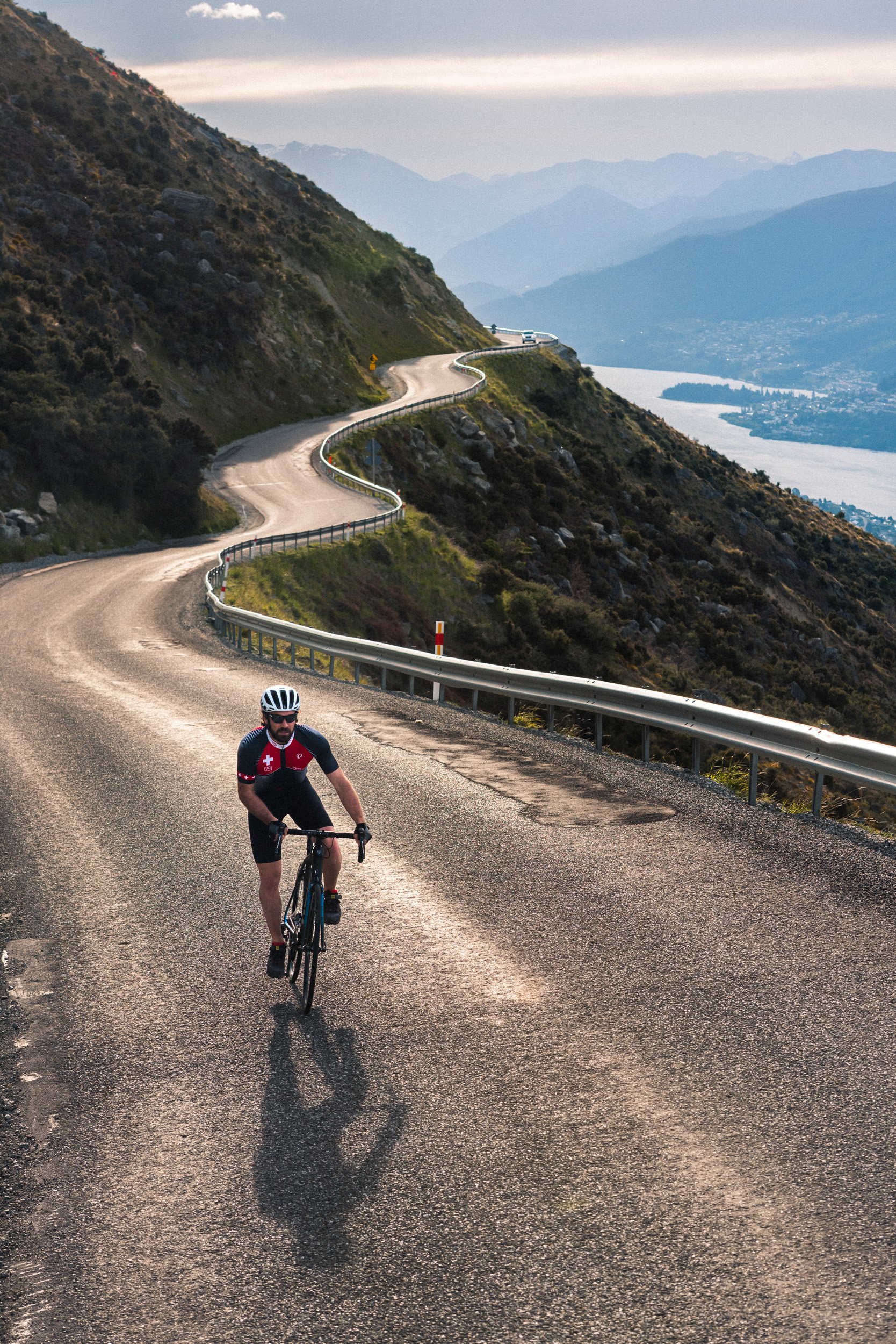 A cyclist riding uphill on a winding mountain road with guardrails, overlooking a lake and distant mountains in the background.