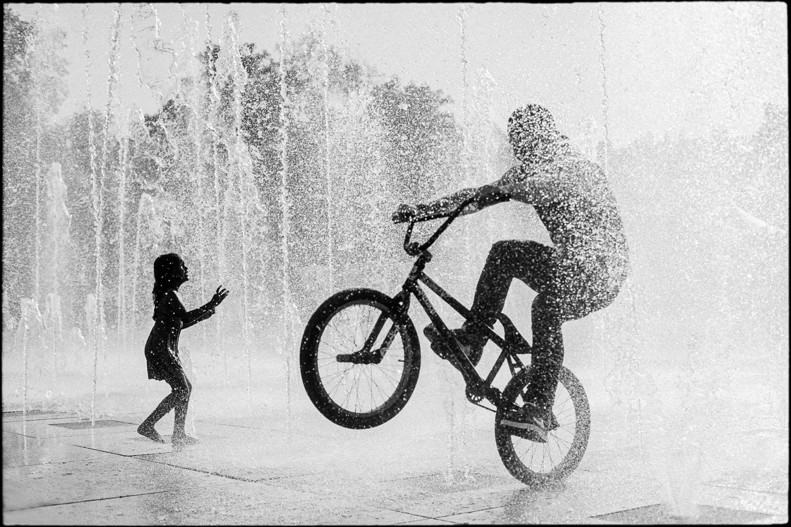 A person riding a bicycle through a water fountain while a little girl plays in front of them.