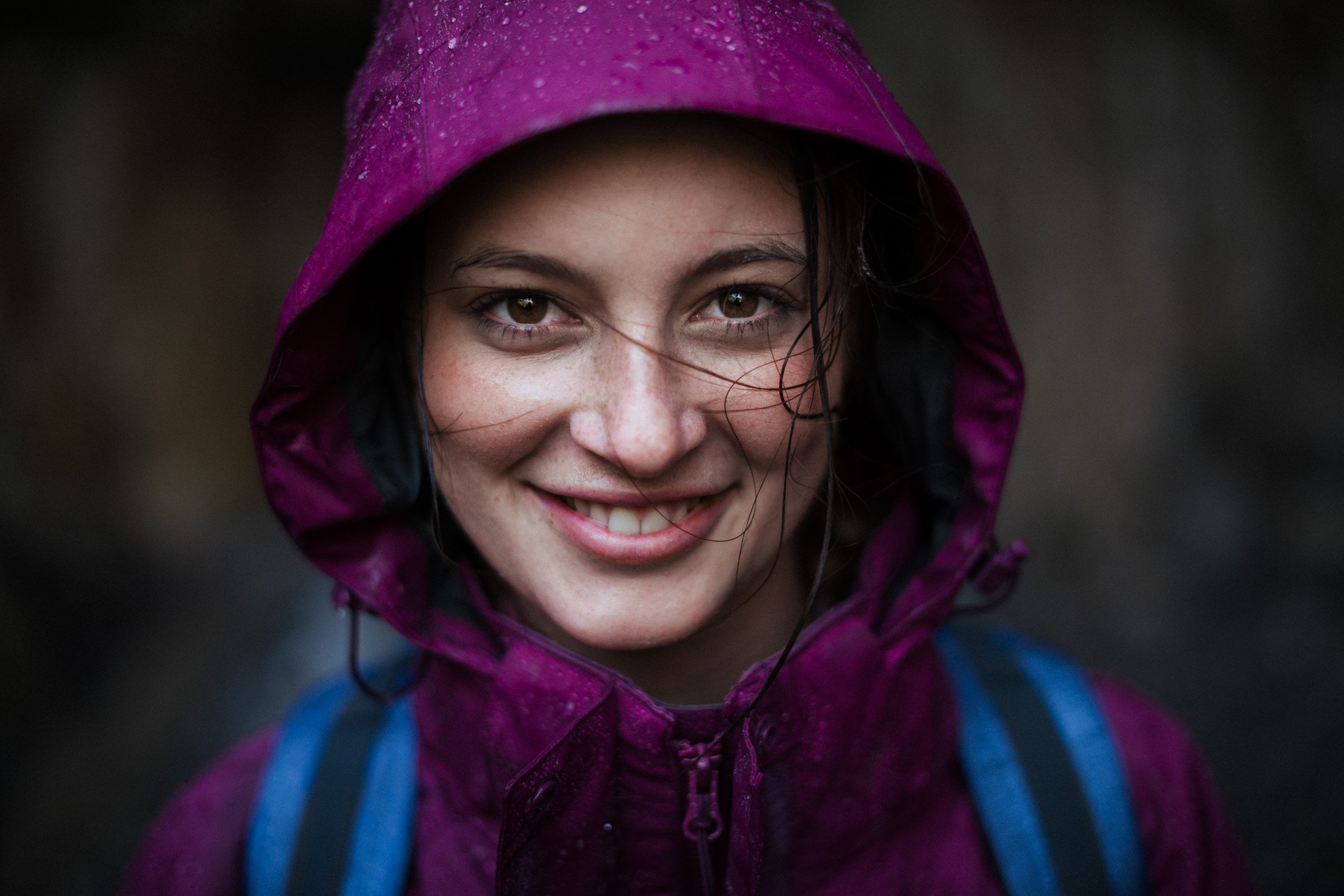 A smiling young woman wearing a purple rain jacket with the hood up, wet from the rain, with a blue backpack visible on her shoulders, outdoors with a dark blurred background.