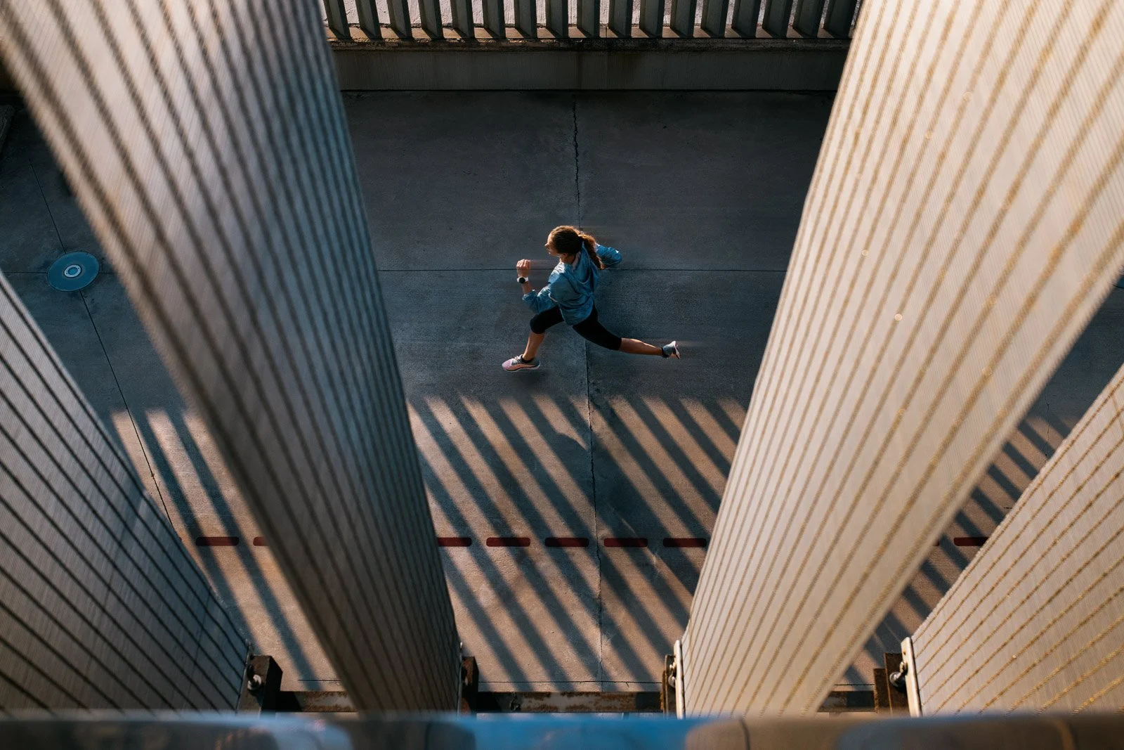 A woman jogging on a concrete sidewalk between two tall vertical buildings, with shadows cast from the buildings creating striped patterns on the ground.