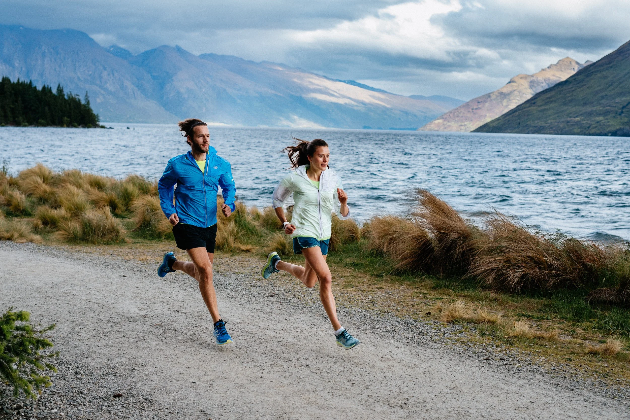 Two people running outdoors along a gravel path next to a lake, with mountains and cloudy sky in the background.