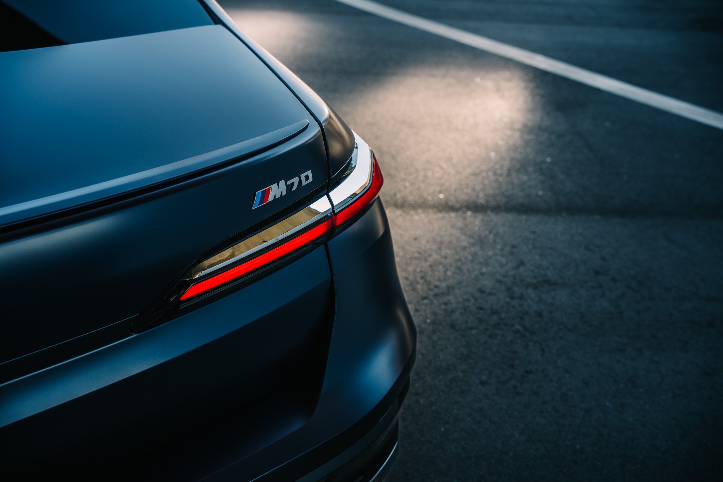 Close-up of the rear end of a dark-colored BMW M770 parked on an empty road with a reflection of sunlight on the surface.