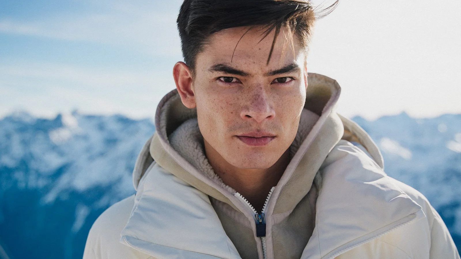 A young man with short dark hair and freckles, wearing a beige jacket and hoodie, standing outdoors in a snowy mountainous landscape.