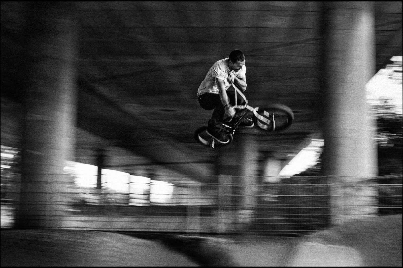 A person performing an aerial trick on a BMX bicycle under a bridge, captured in black and white motion blur.