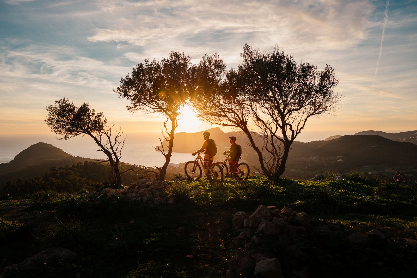 Two people riding bicycles on a trail during sunset, with trees on either side and mountains in the background.