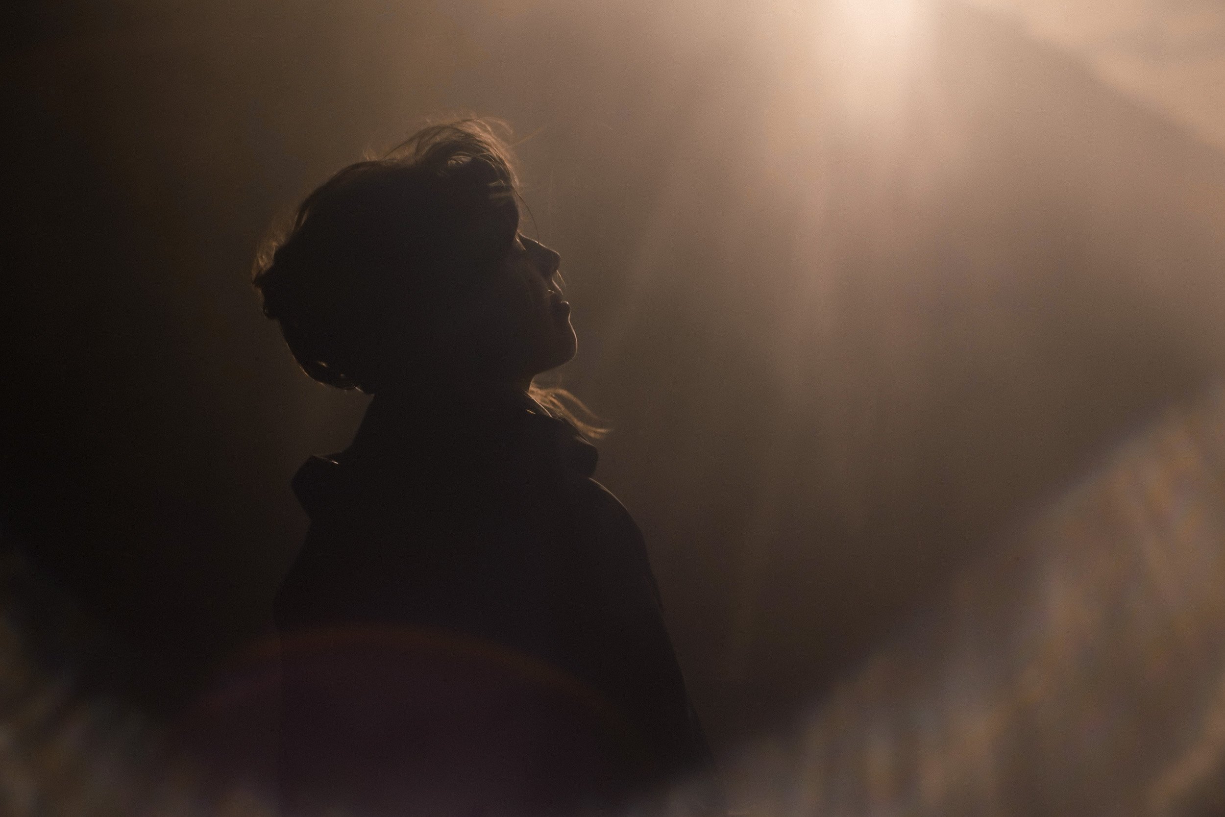 Silhouette of a woman with slightly wavy hair, backlit with warm light, creating a shadowy profile against a dark background.