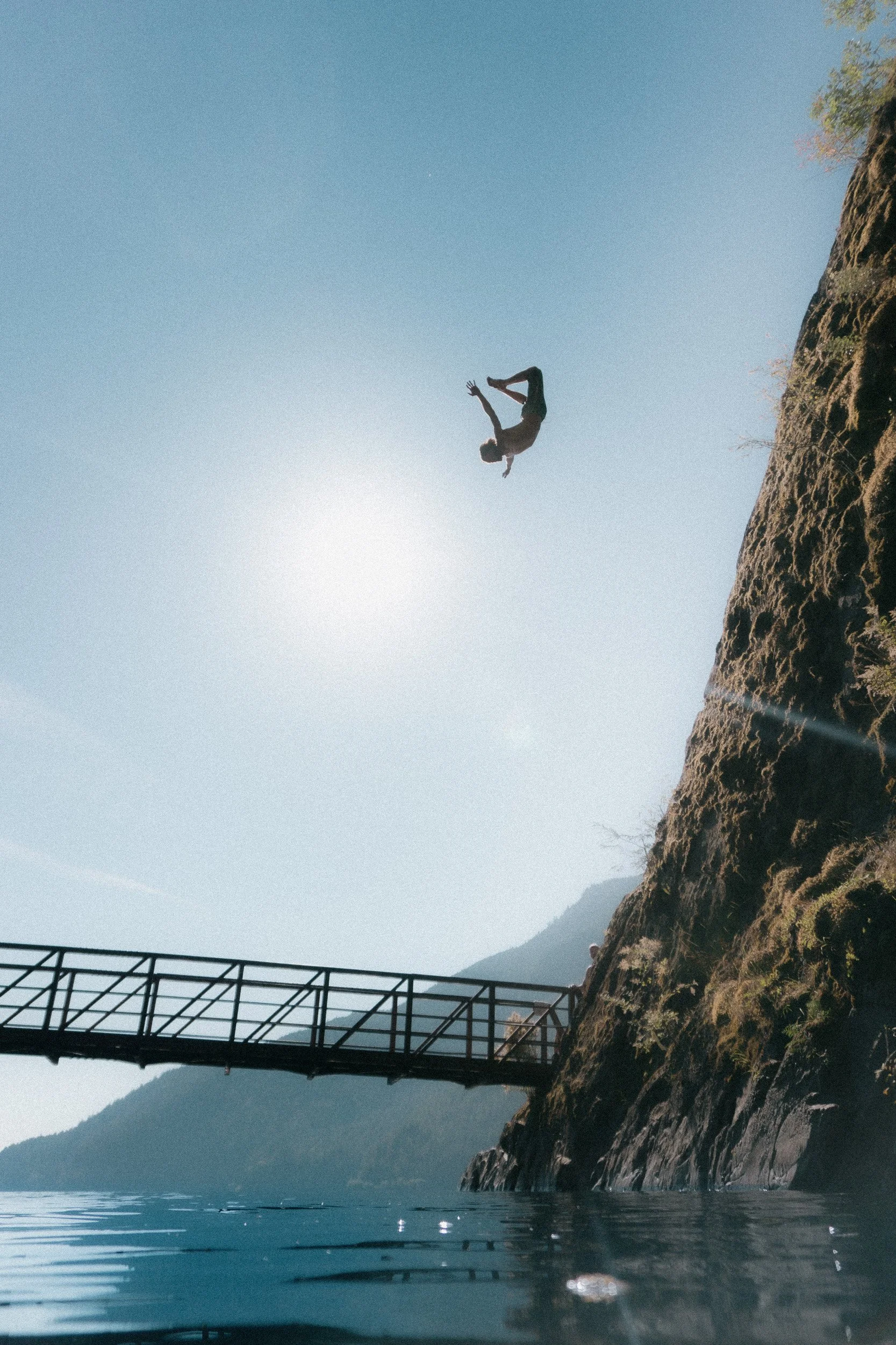 Person mid-air diving into water near a rocky cliff and bridge, with mountains and clear sky in the background.