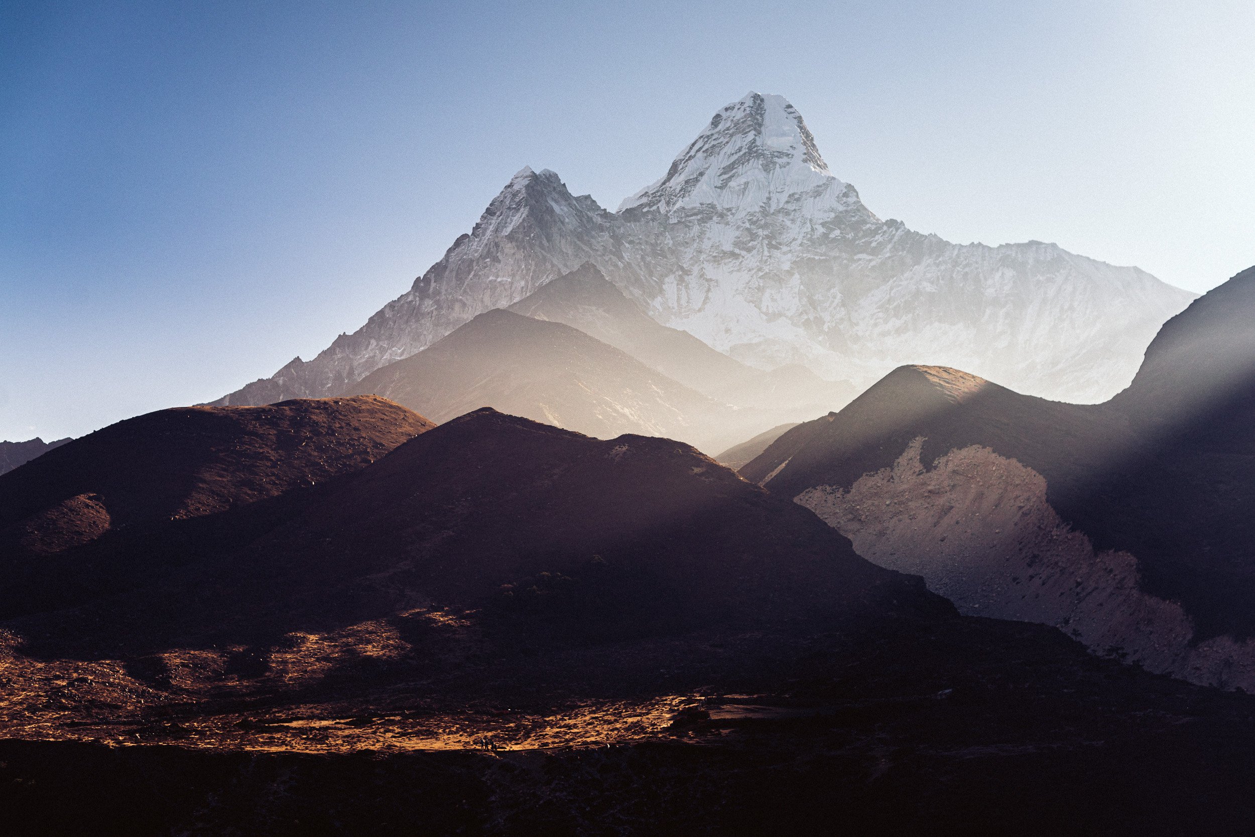 Snow-capped mountain peak behind brown rolling hills with clear sky.