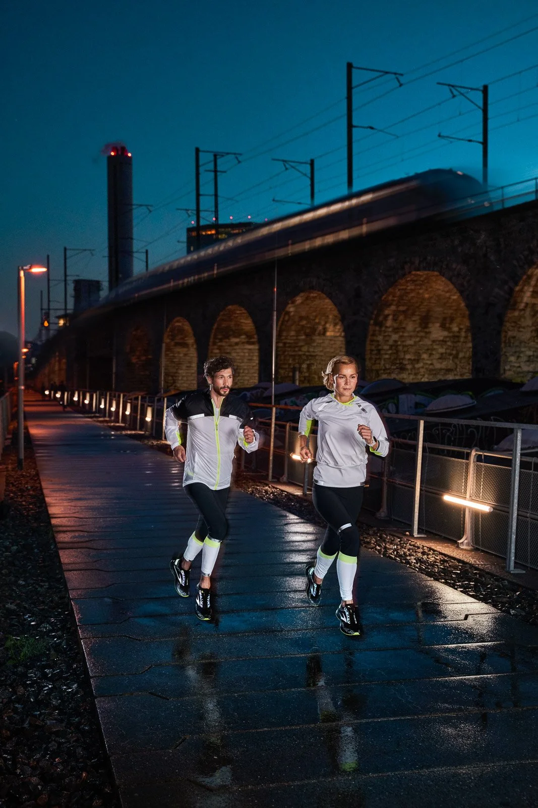 Two people running outdoors at night with wet pavement, illuminated by streetlights, train tracks, and a cityscape in the background.
