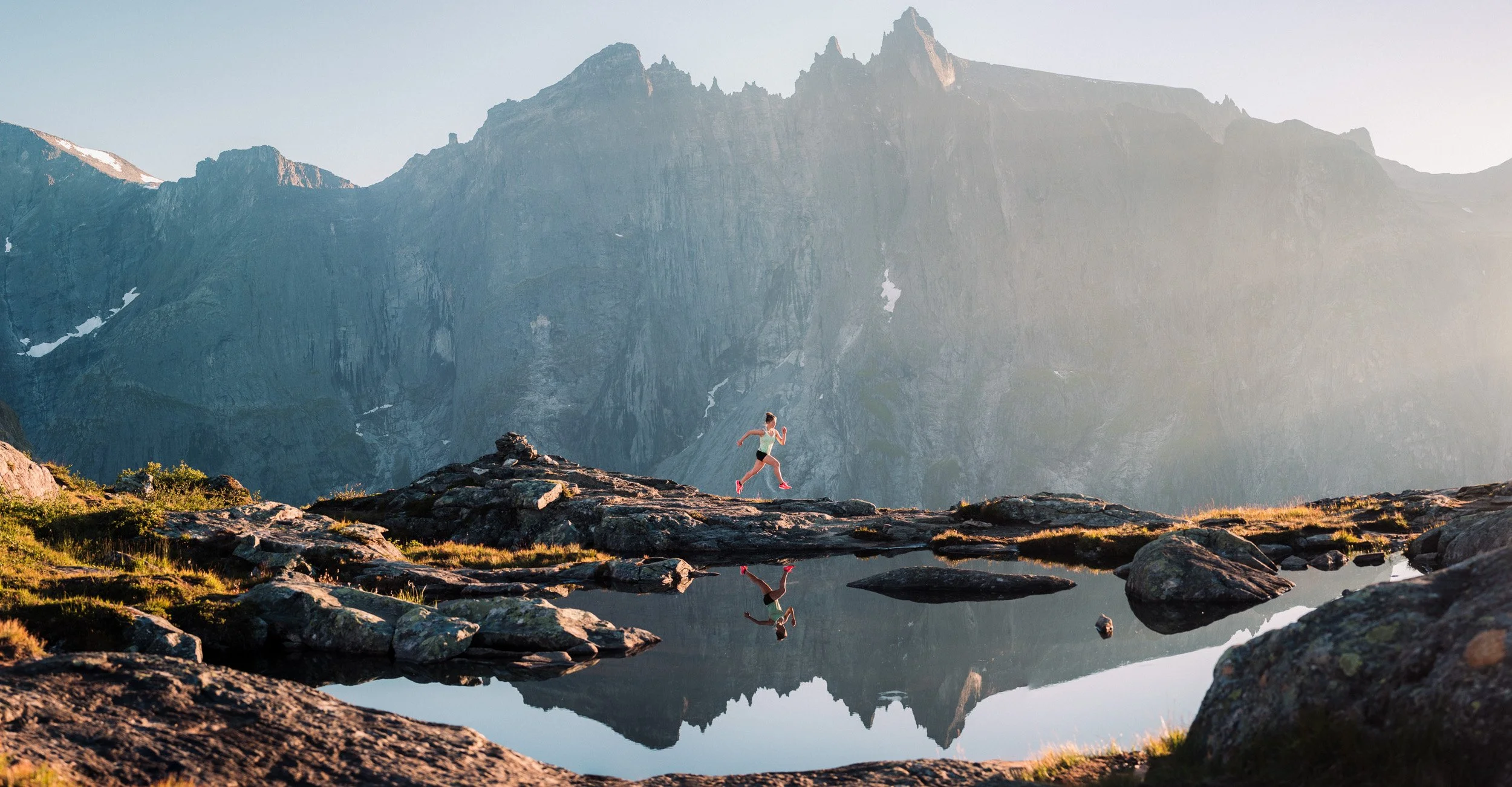 A person running on rocky terrain near a reflective body of water with mountains in the background