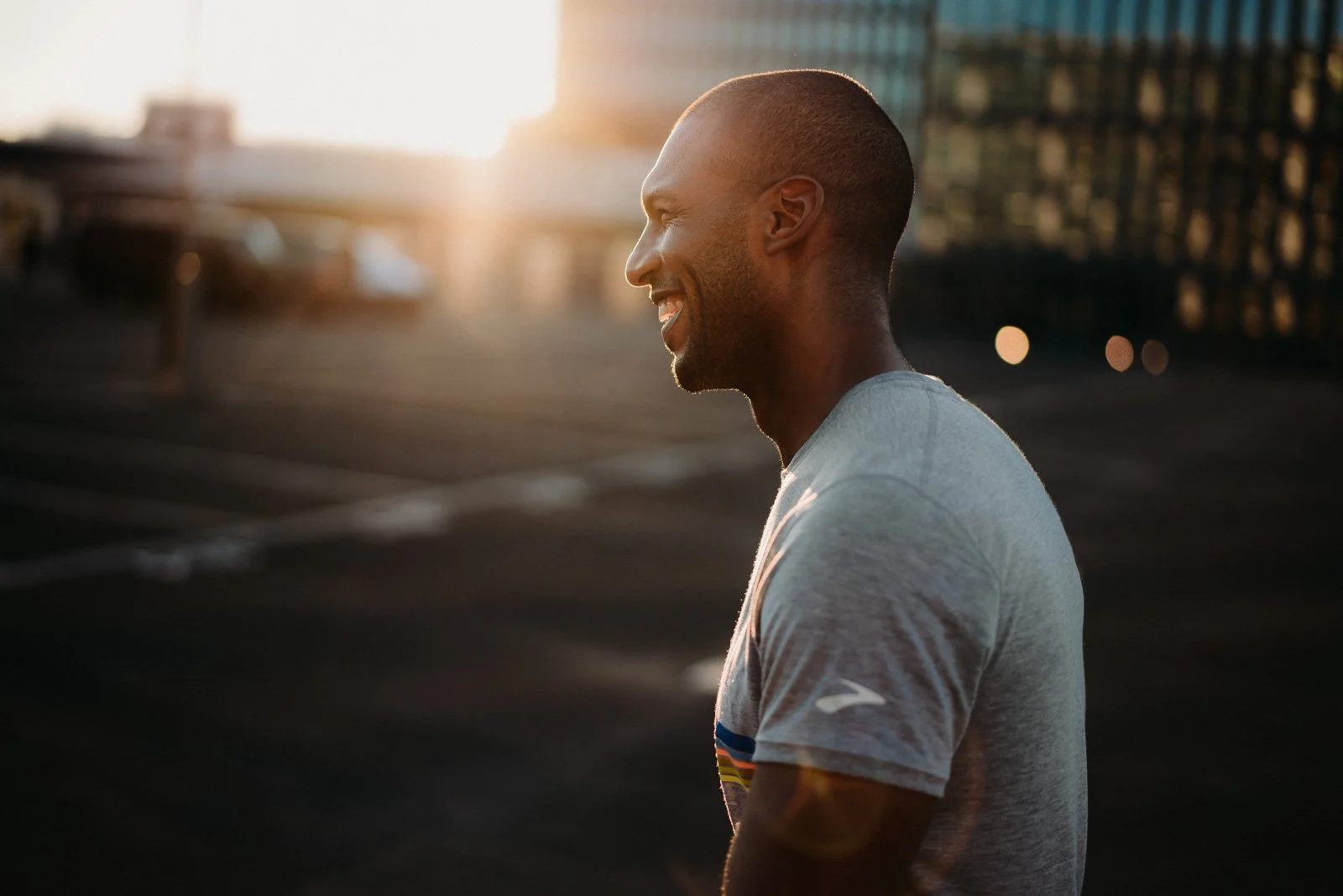 Side profile of a smiling man wearing a gray T-shirt outdoors during sunset with city buildings in the background.