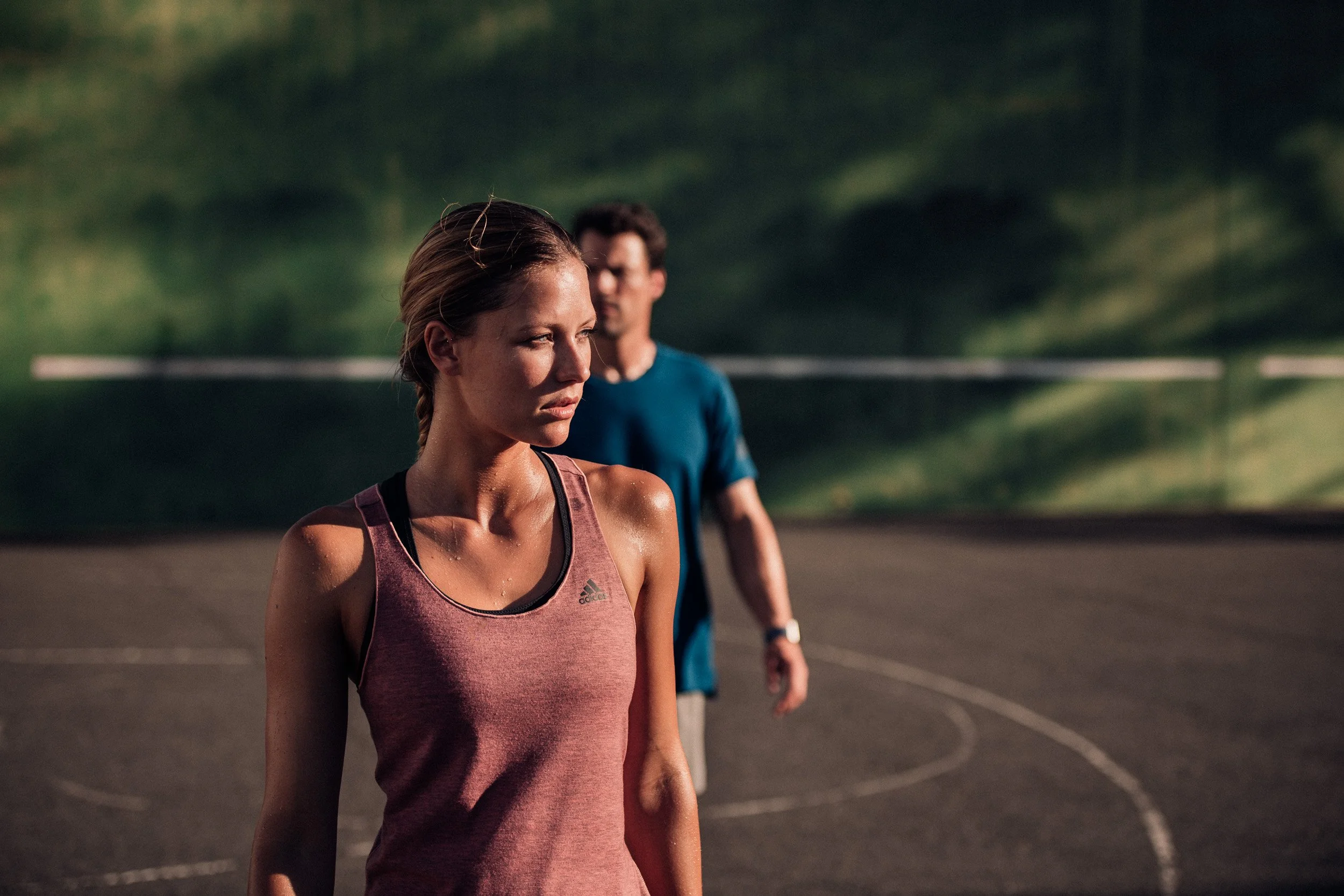 A woman in athletic wear standing on a basketball court with a man in the background during sunset.
