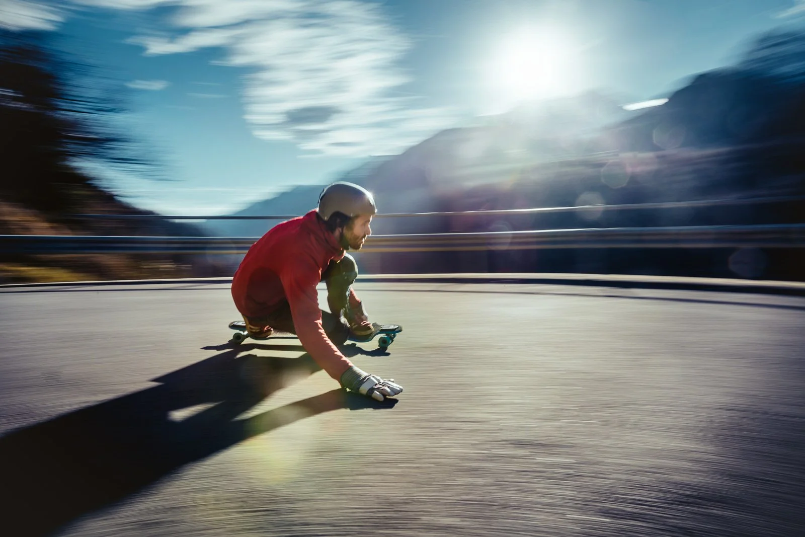 A person wearing a helmet and red jacket is skateboarding downhill on a curved road during daytime, with mountains and sky in the background.
