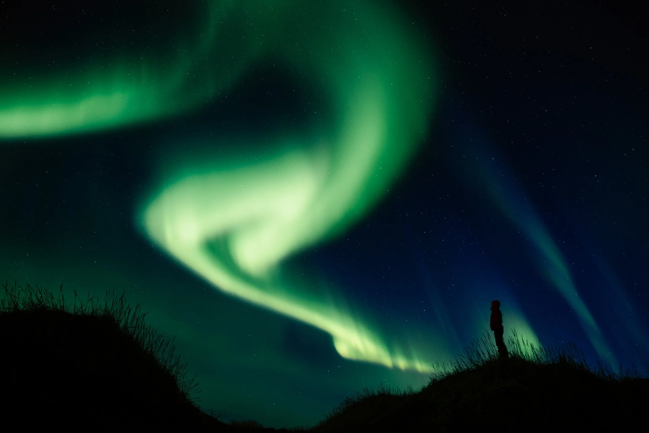 A person standing on a hill under the night sky illuminated by green and blue Northern Lights, with stars visible.