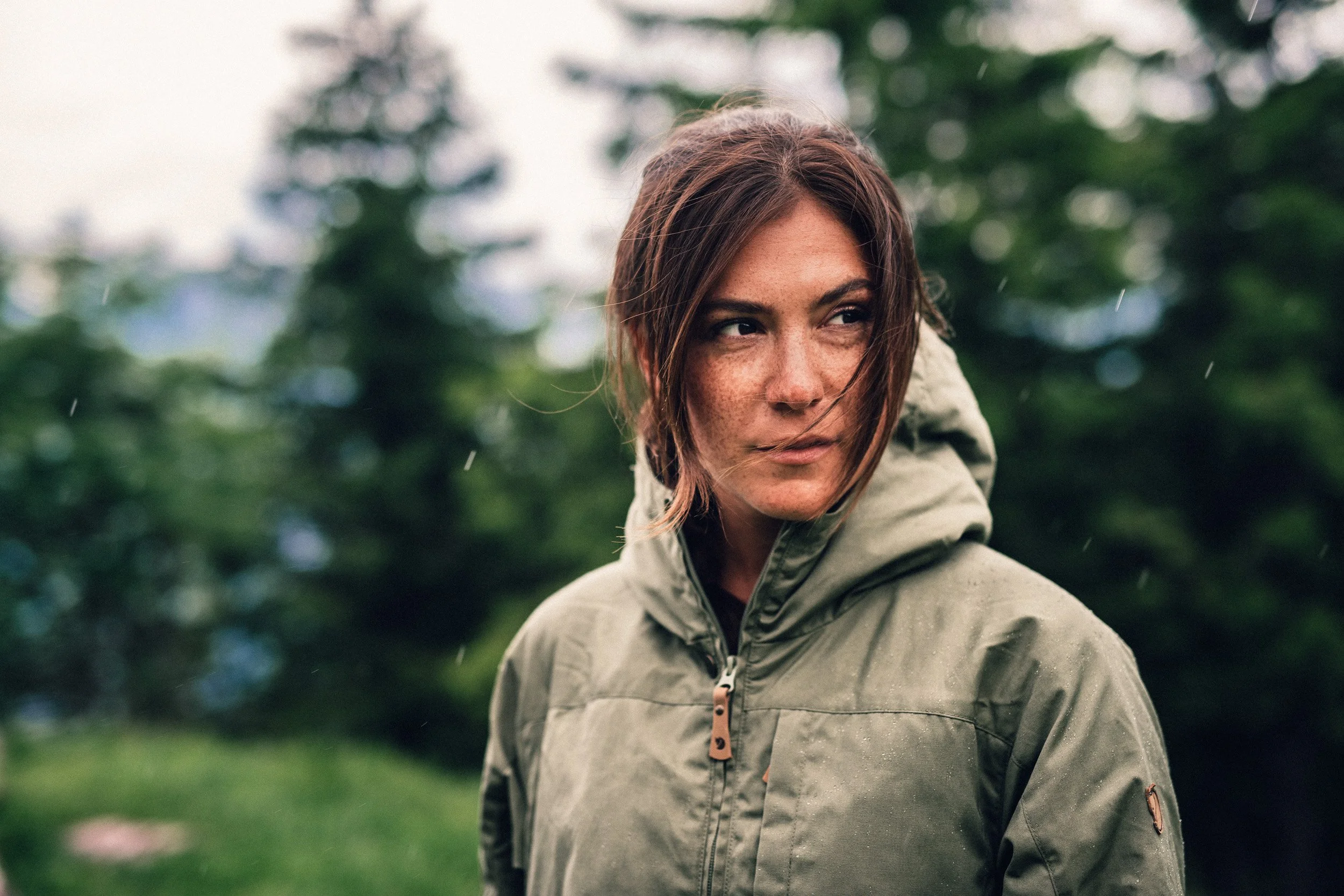 A woman with brown hair and freckles wearing a green rain jacket outdoors in a forest with blurred trees in the background.