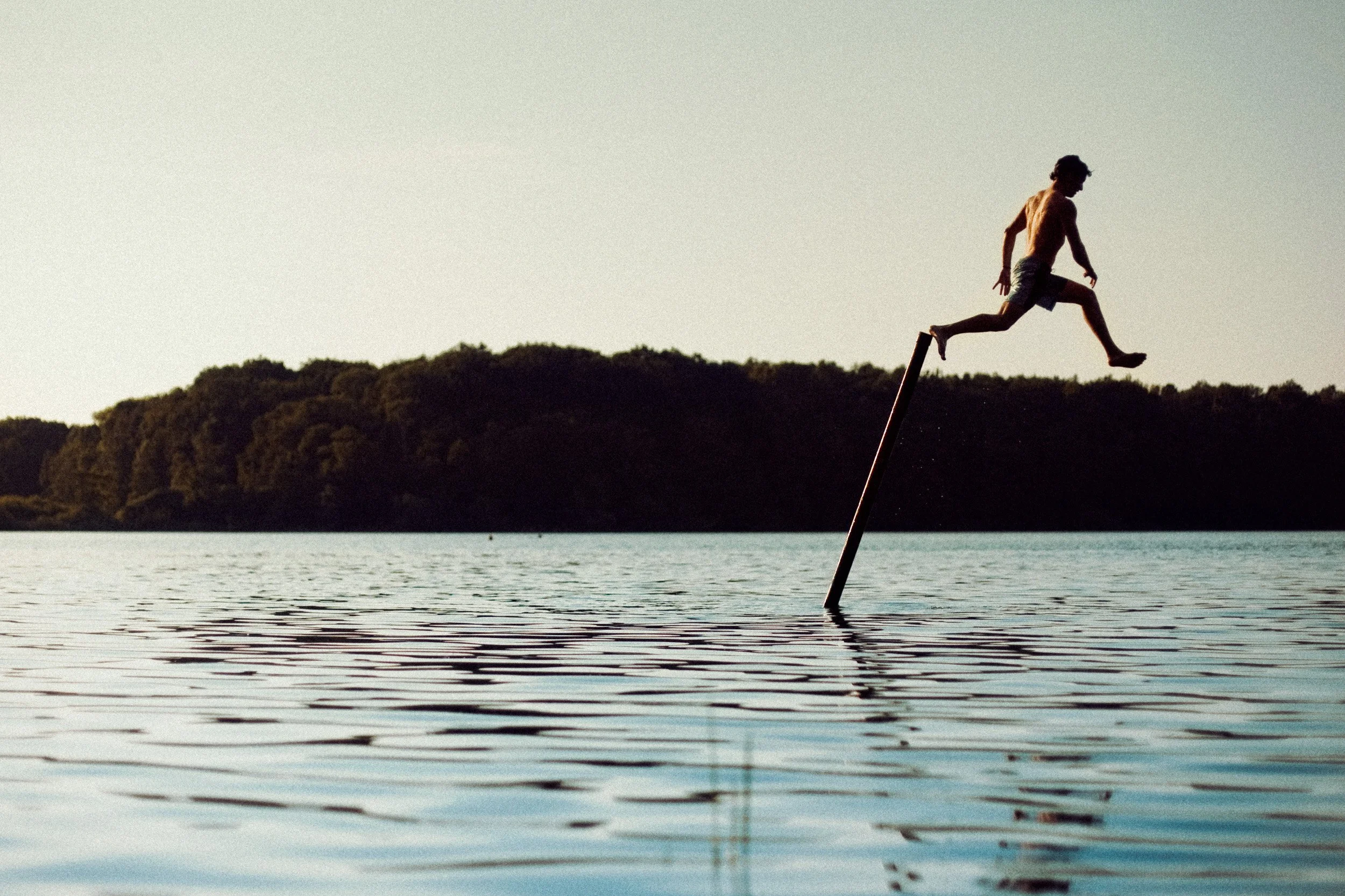 A person jumping off a stick protruding from the water onto a floating log, with a background of trees and a clear sky.