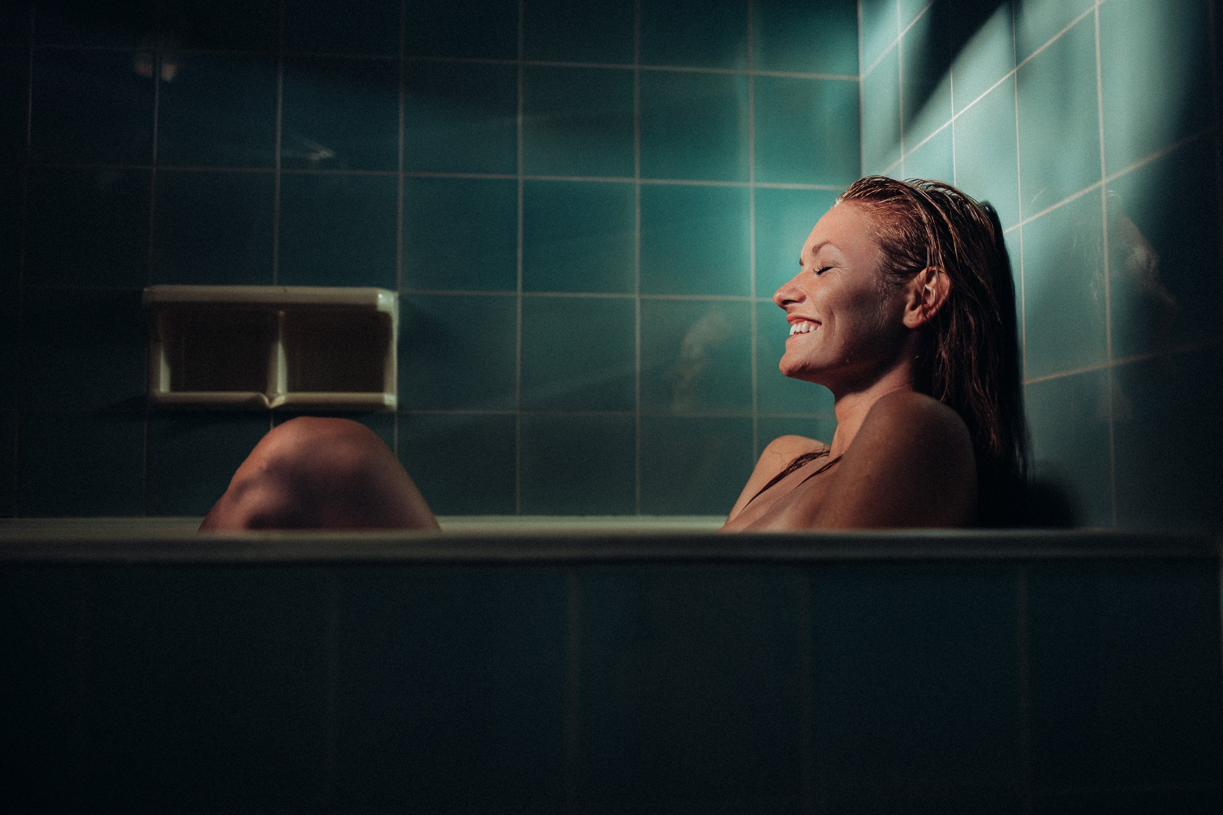 A woman relaxing in a bathtub with her eyes closed and smiling, in a tiled bathroom with greenish-blue tiles.