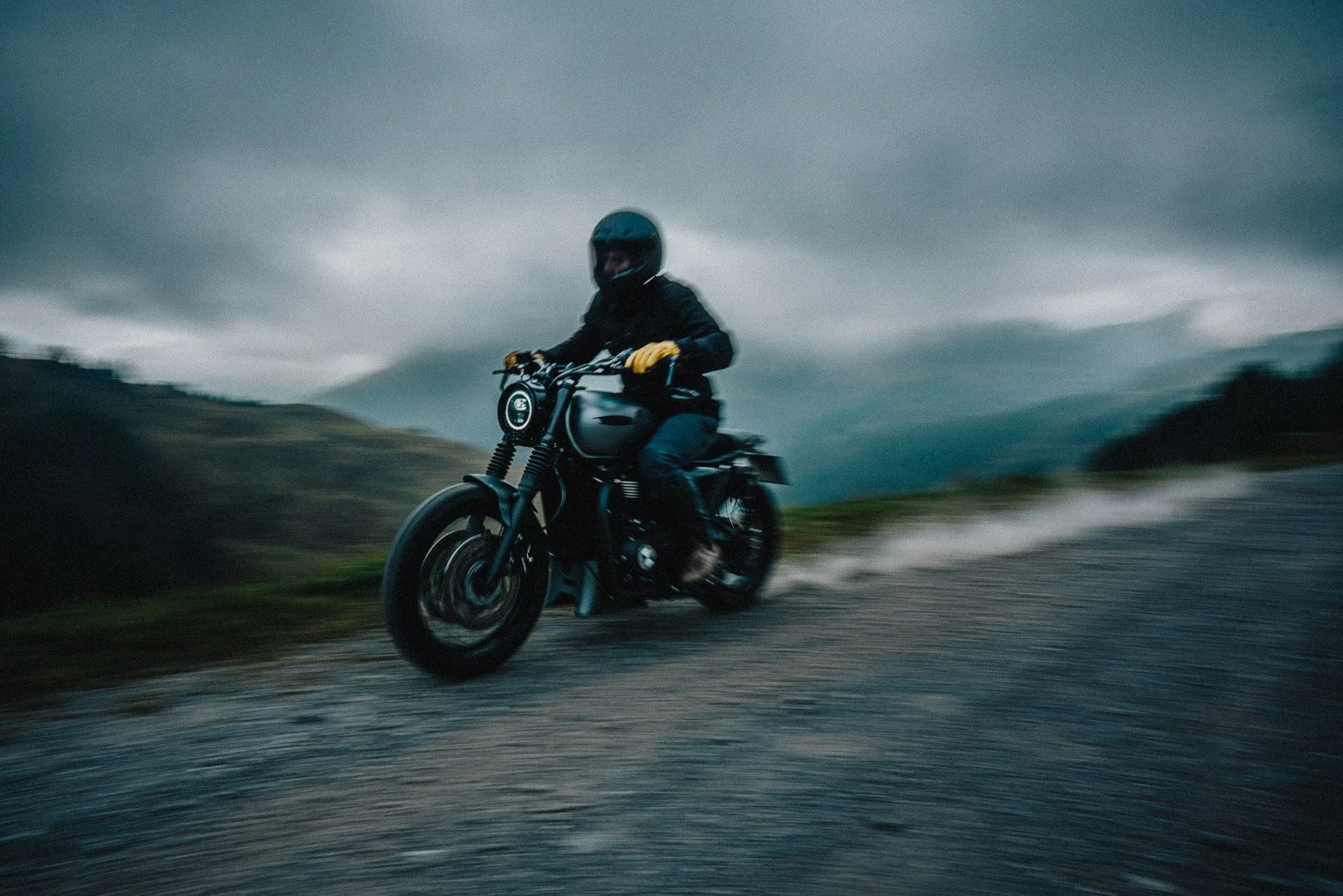 A person riding a black motorcycle on a gravel road in a mountainous landscape under a cloudy sky, wearing a helmet and glove.