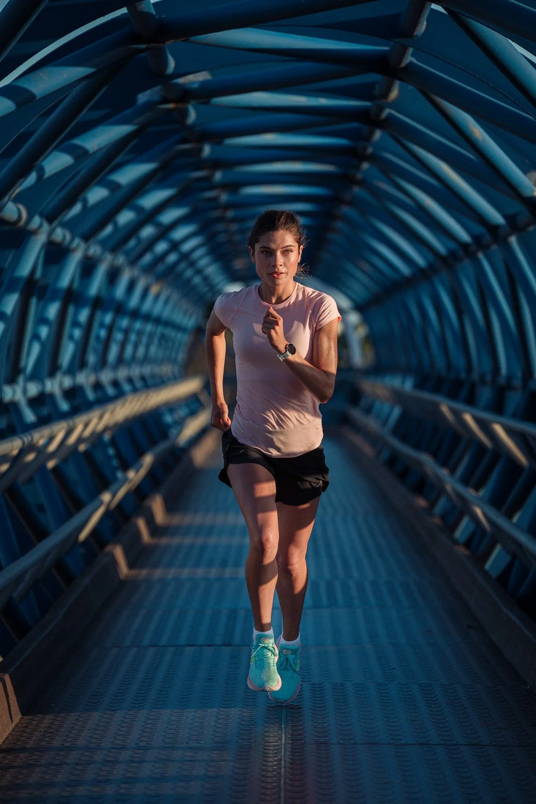 A woman running through a blue steel tunnel, wearing a light pink shirt, black shorts, and turquoise running shoes.