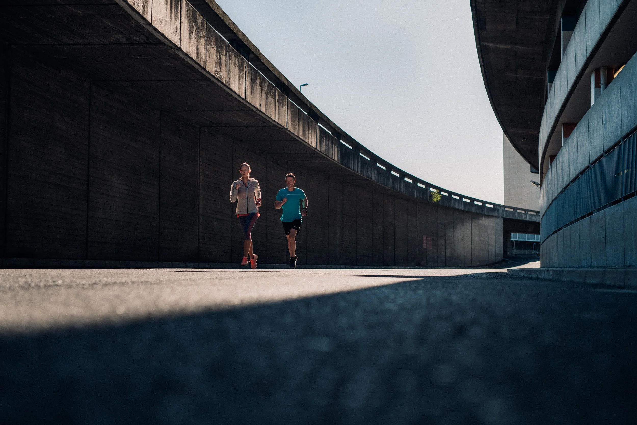 Two people jogging under a curved overpass in an urban setting.