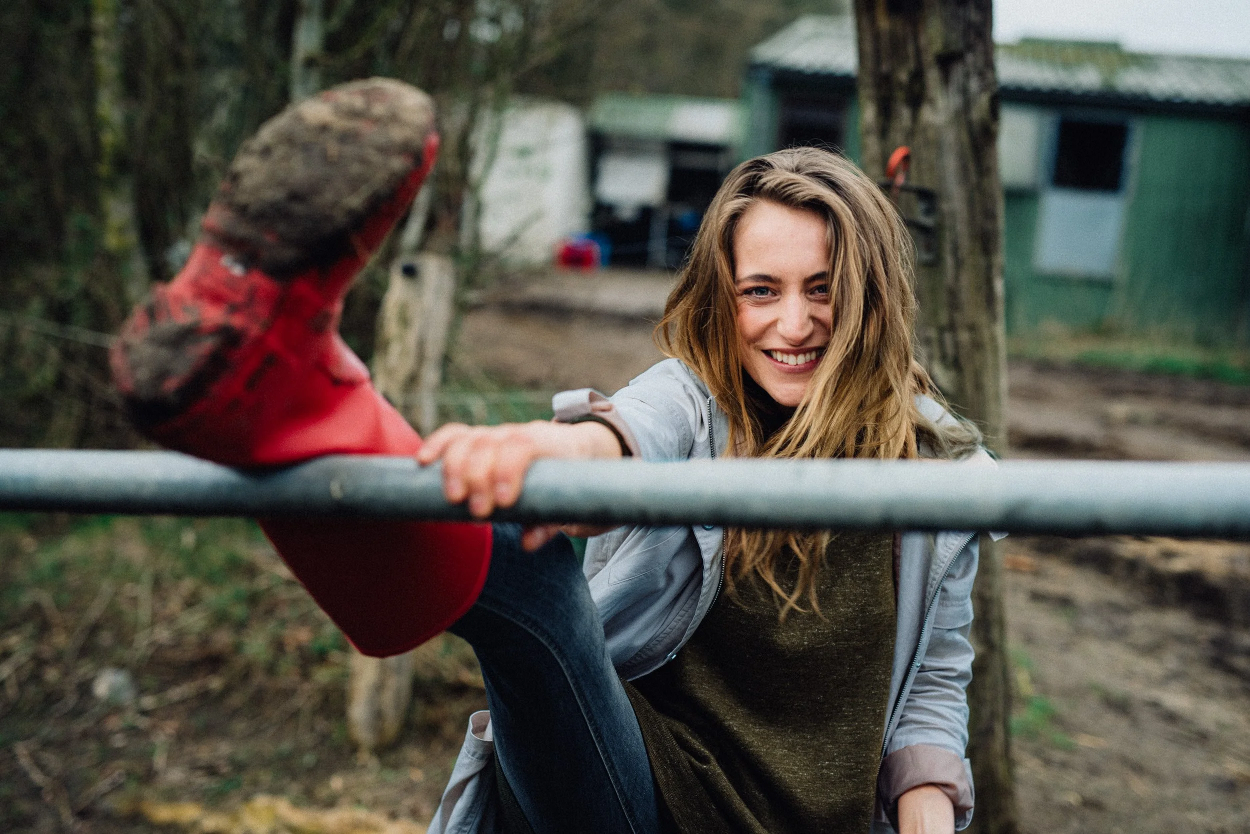 A young woman outdoors, smiling and extending her leg with a red boot resting on a metal bar.