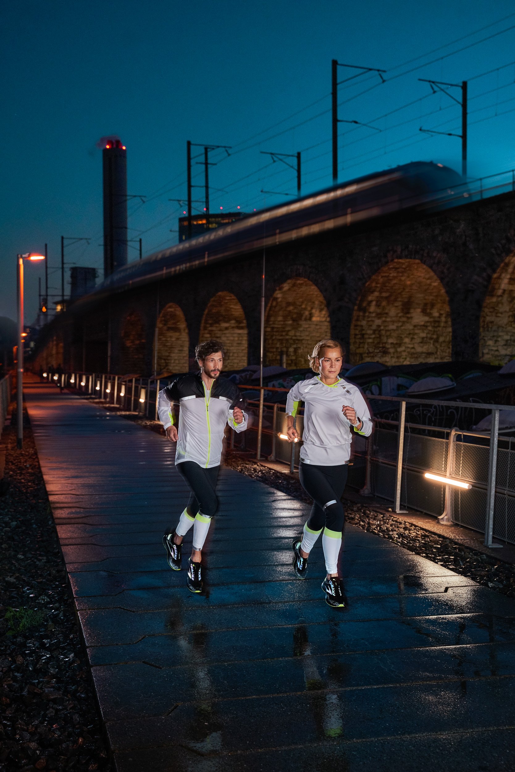 Two people running at night on a wet sidewalk near a raised train track with a cityscape in the background.