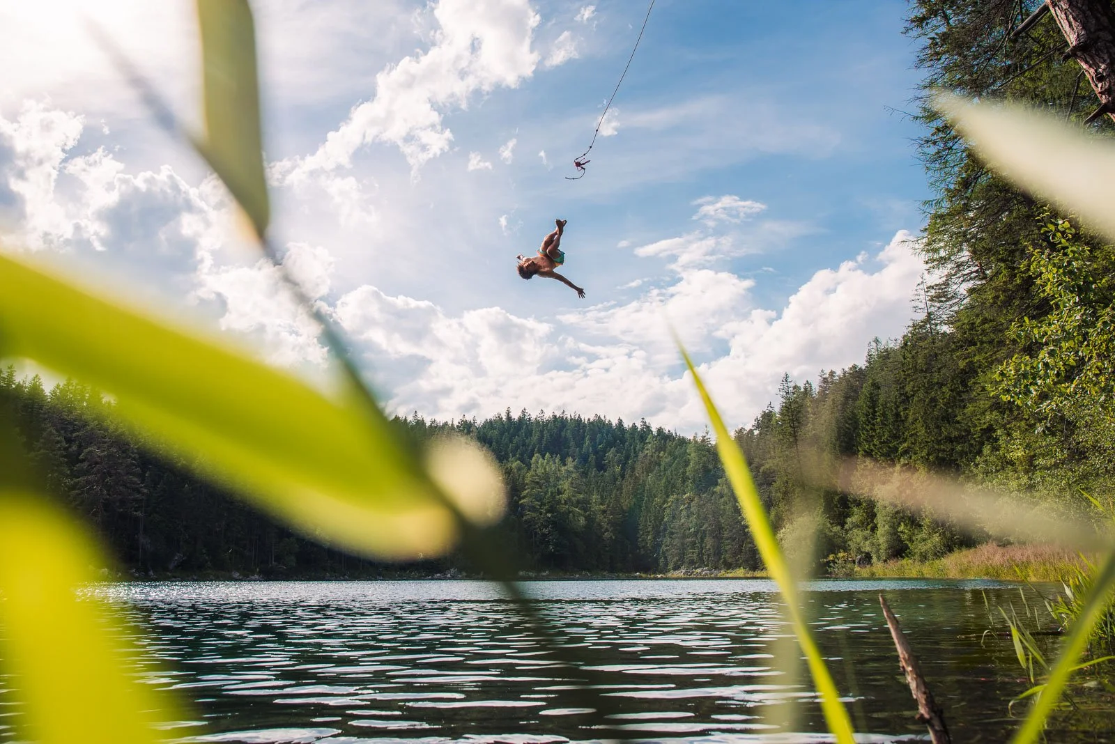 Young person mid-air jumping off a rope swing over a lake, surrounded by trees and a cloudy sky.