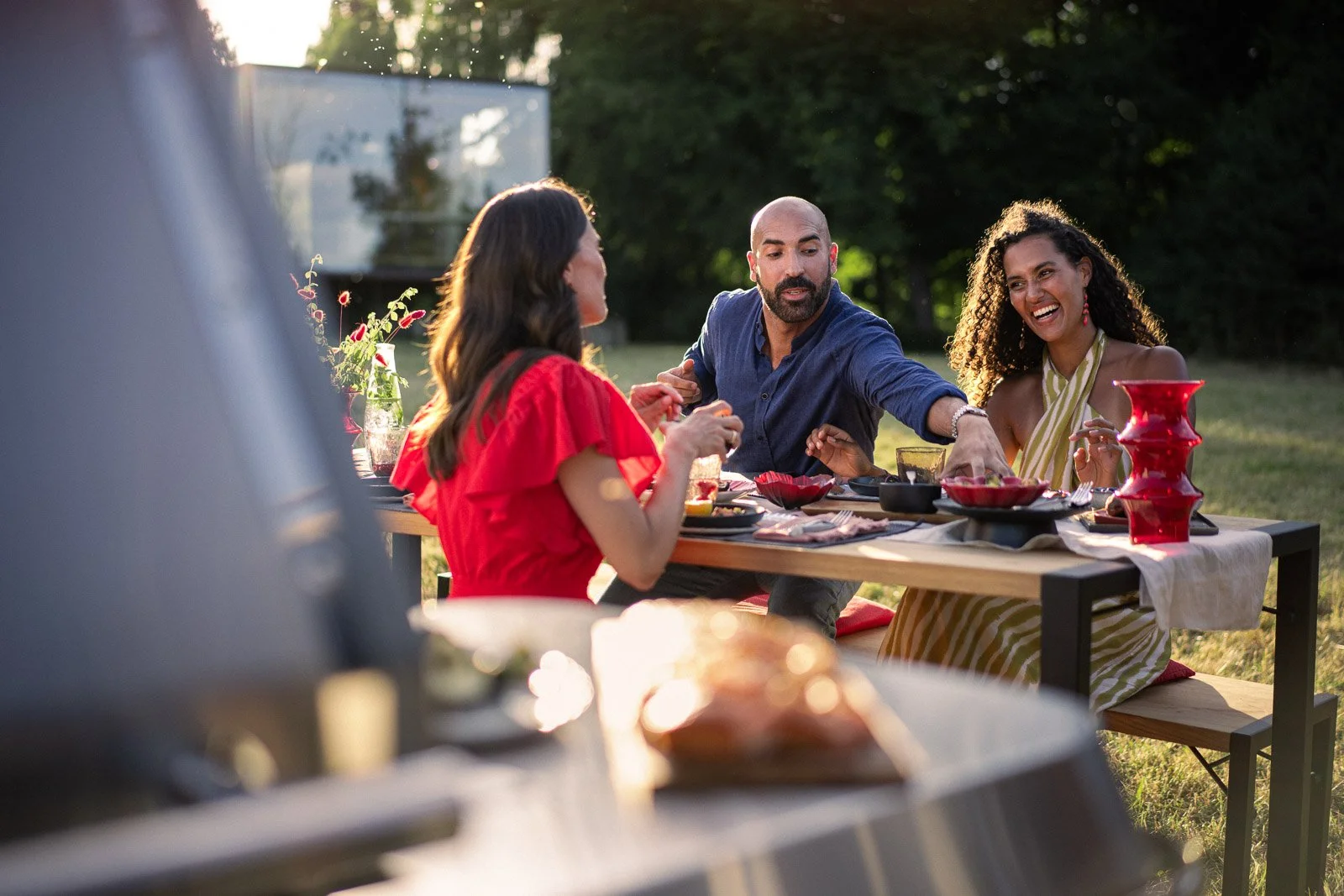 Three people sitting at a table outdoors, enjoying a meal together during the evening. The scene depicts a diverse group, with one woman wearing a red dress, a man in a blue shirt, and another woman in a striped dress. They are smiling and appear to 