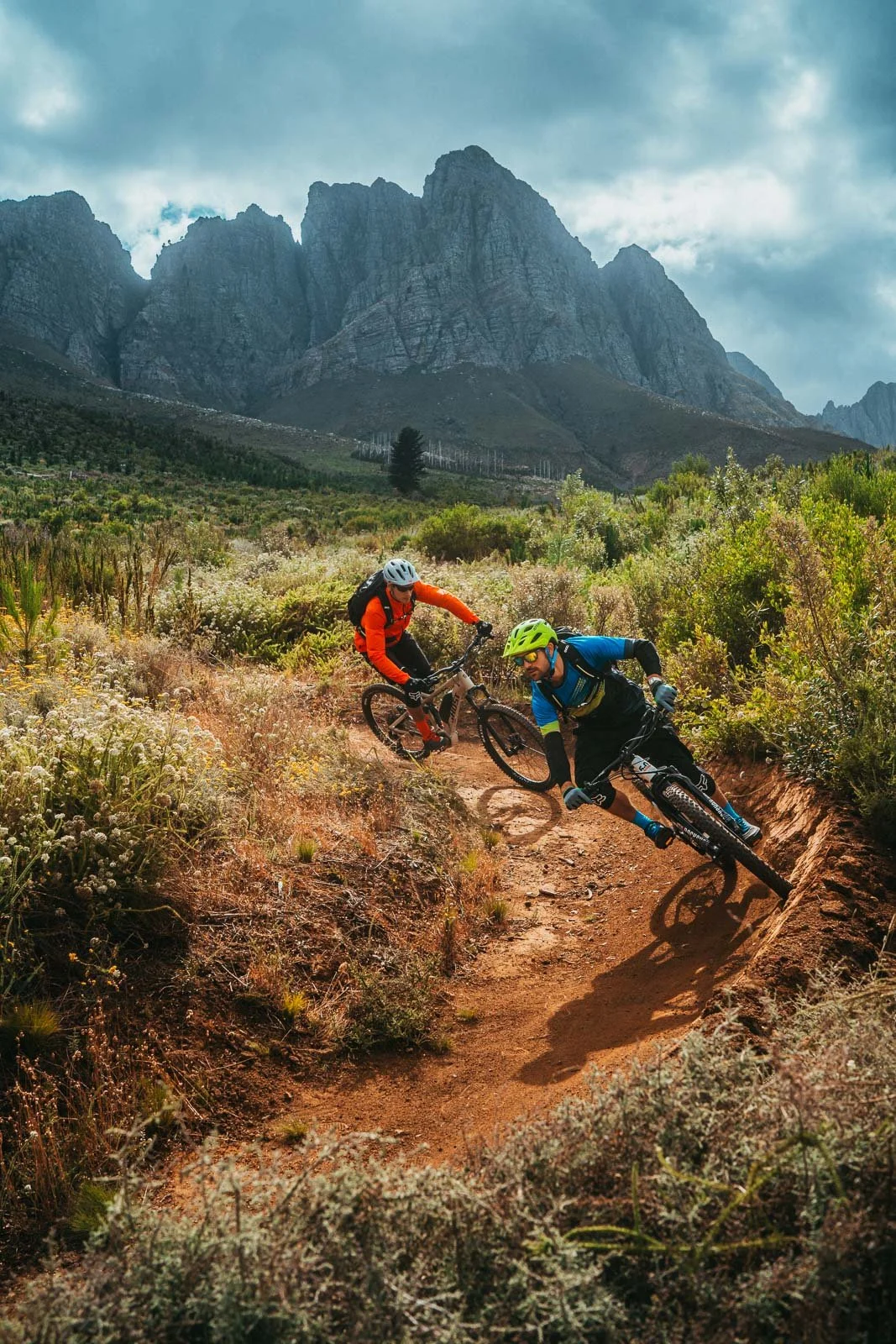 Two mountain bikers riding on a dirt trail through a rugged landscape with tall mountains in the background.