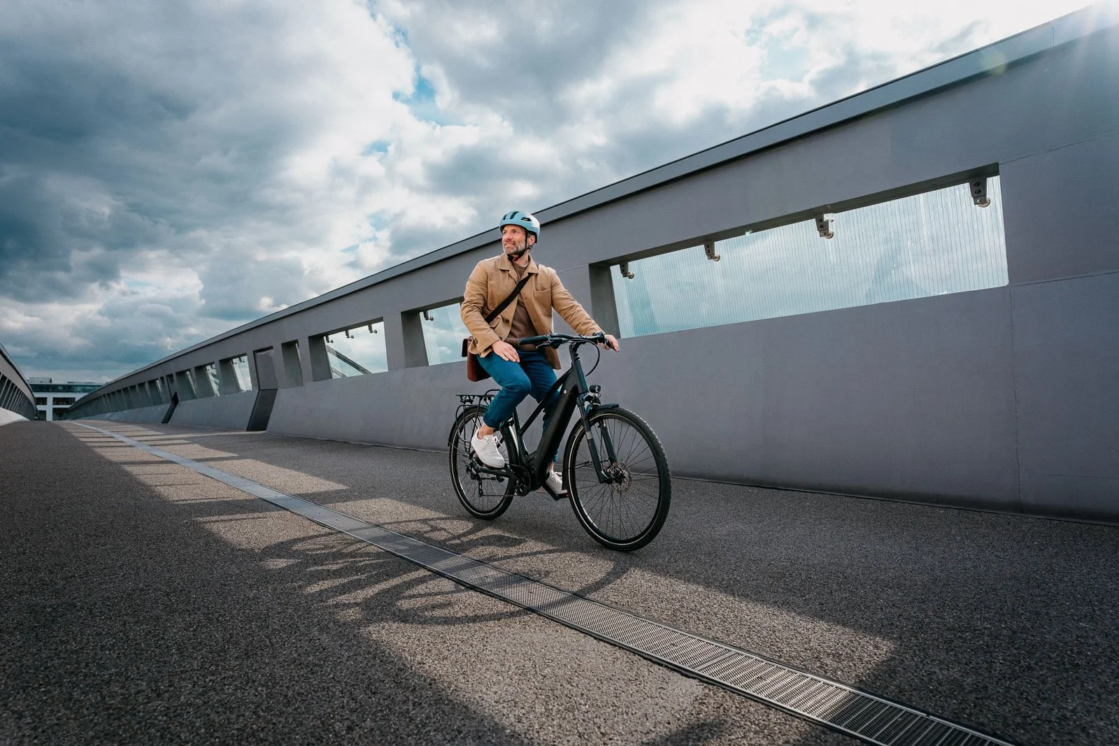 A man riding a black electric bicycle on a modern, curved bridge with glass-paneled safety barriers under a cloudy sky.