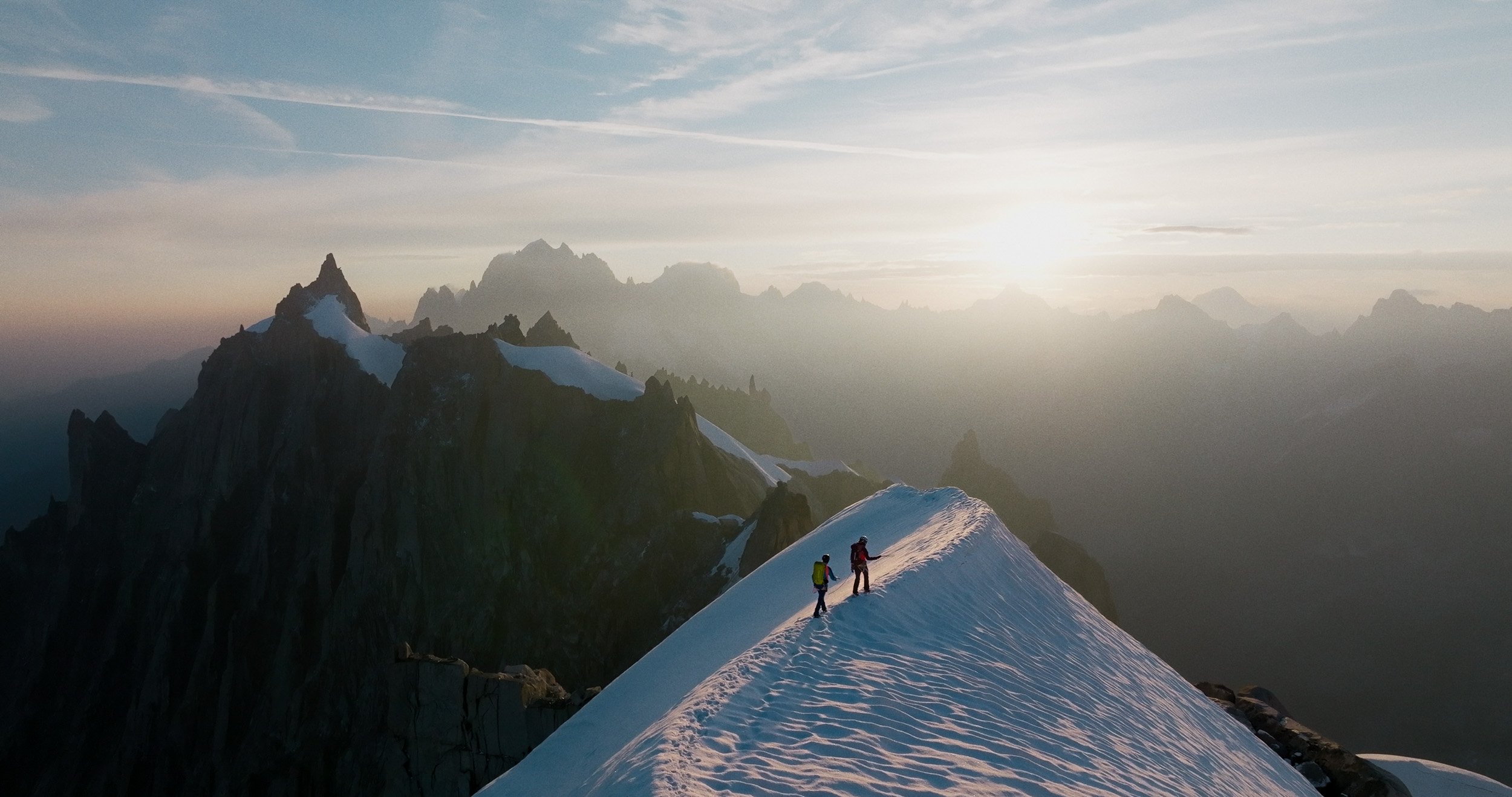 Two mountain climbers on a snowy ridge at sunset, with rugged mountains in the background and a partly cloudy sky.