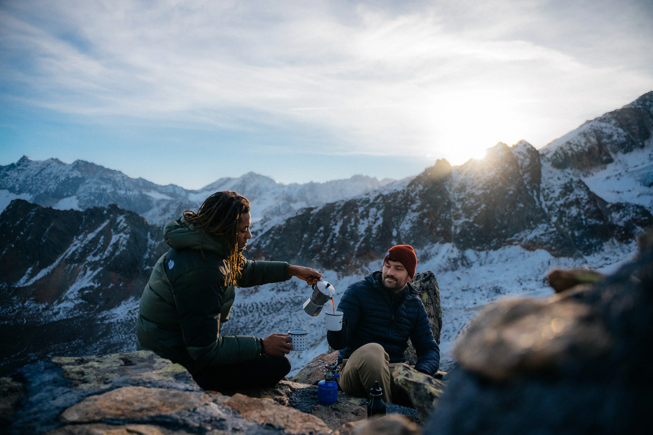 Two people camping on rocky terrain in a mountain range during sunset. One person is pouring a beverage from a coffee pot into a mug held by the other person.