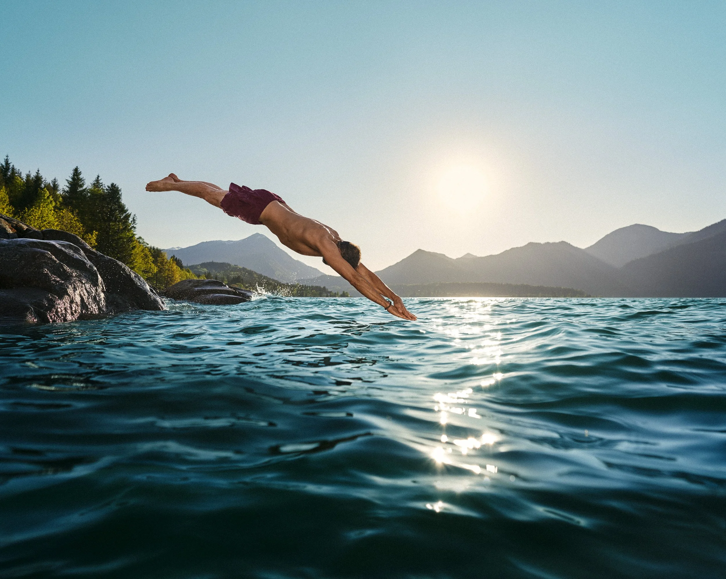 A man diving into a lake with mountains and trees in the background during sunset.
