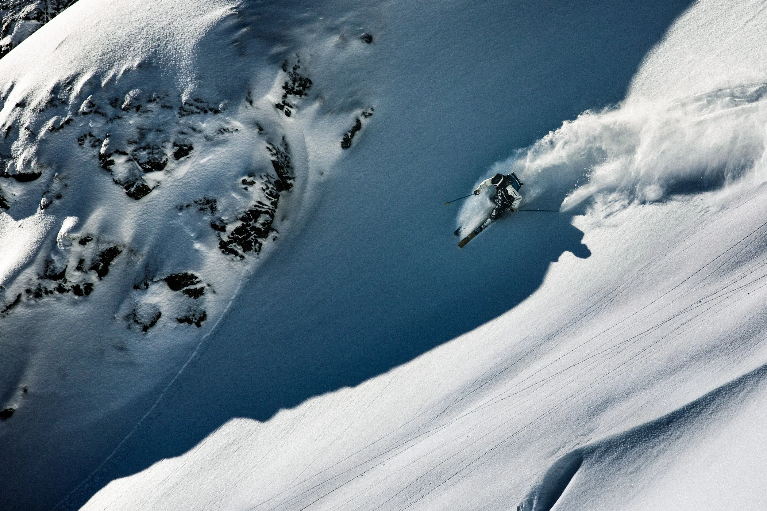 A skier in black and white gear navigating a steep snowy mountain slope with rocks and snow crashing behind him.