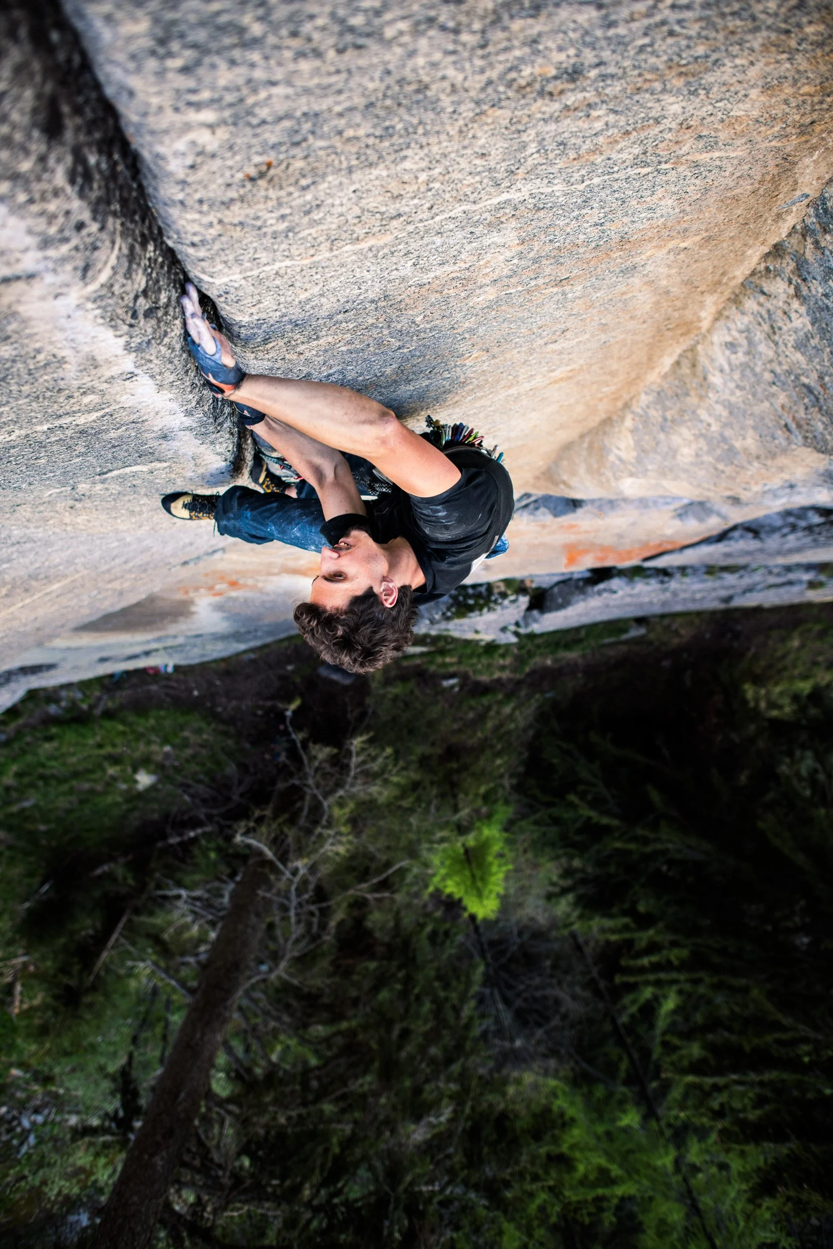 A man climbing a tall rock wall outdoors, wearing climbing shoes and black clothing, surrounded by green trees.