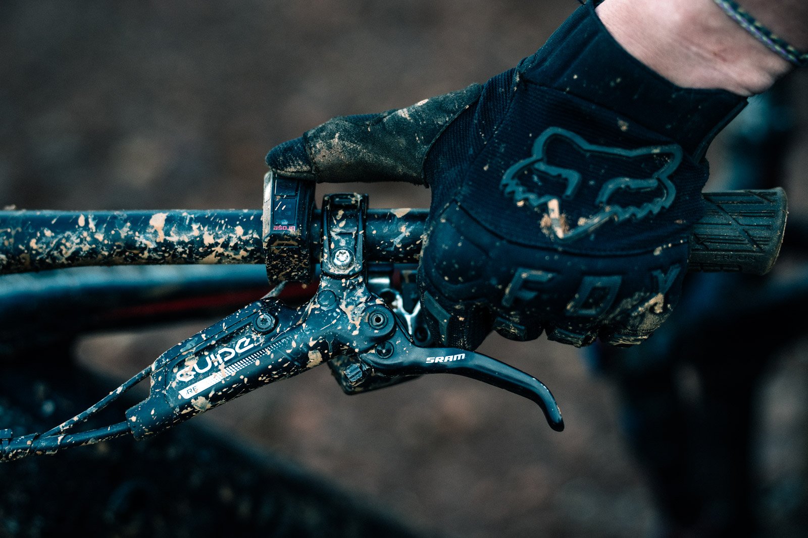 Close-up of a mountain bike handlebar covered in mud, with a gloved hand gripping the right side of the handlebar.