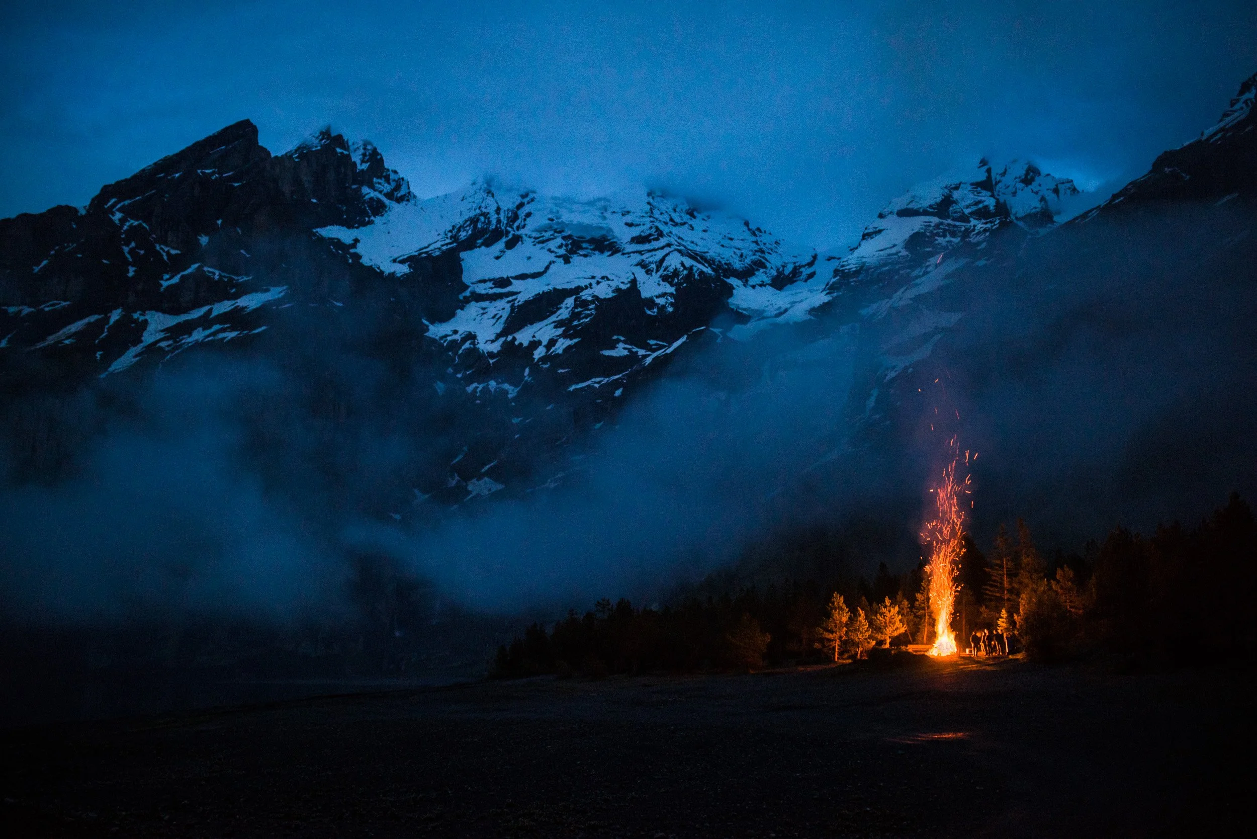 Snow-capped mountains at dusk with a small campfire and trees in the foreground.