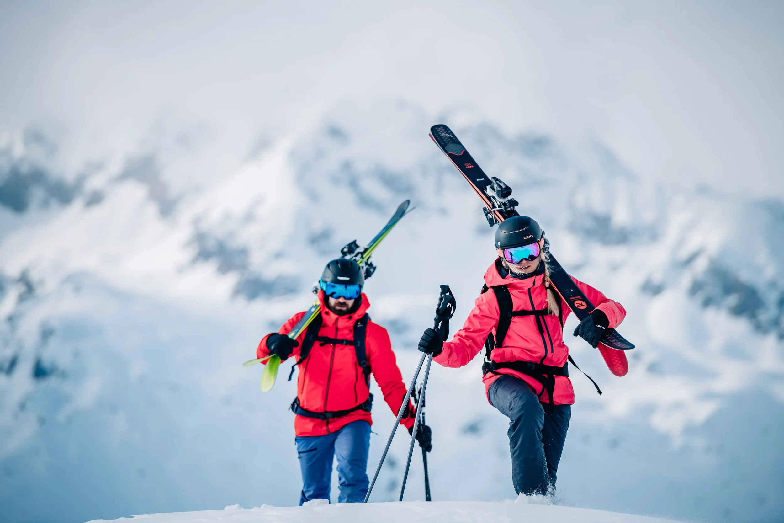 Two skiers wearing red jackets and helmets carrying skis in a snowy mountain landscape.