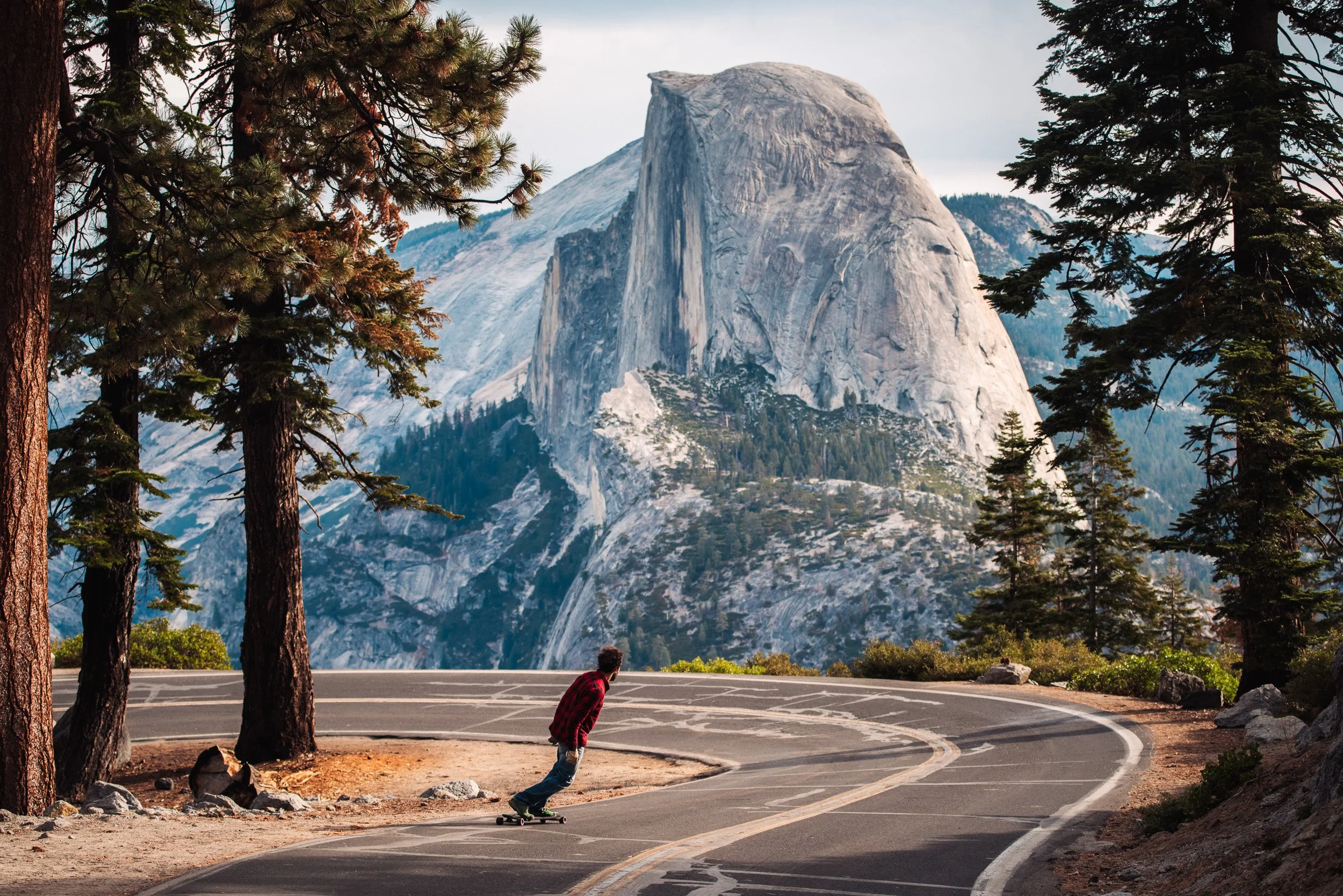 A boy skateboarding on a curving road surrounded by pine trees, with a large granite mountain in the background.