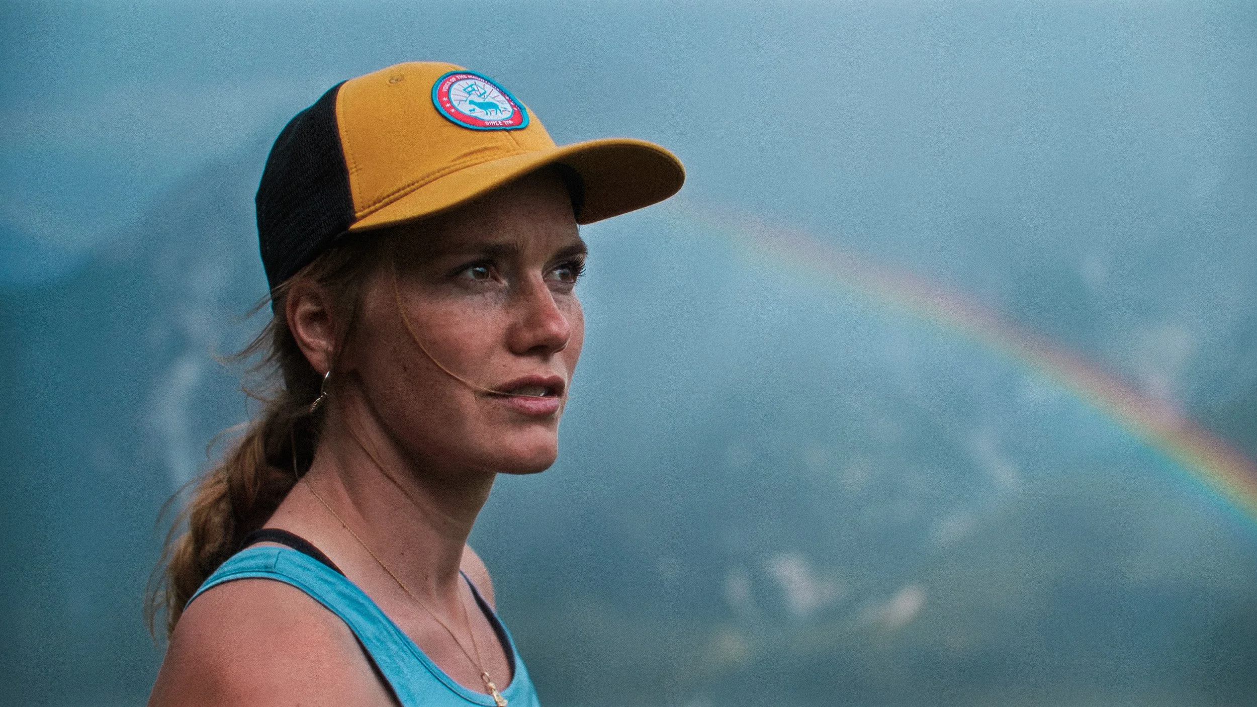 A woman wearing a yellow and black cap and a blue tank top, with a rainbow in the cloudy sky behind her.