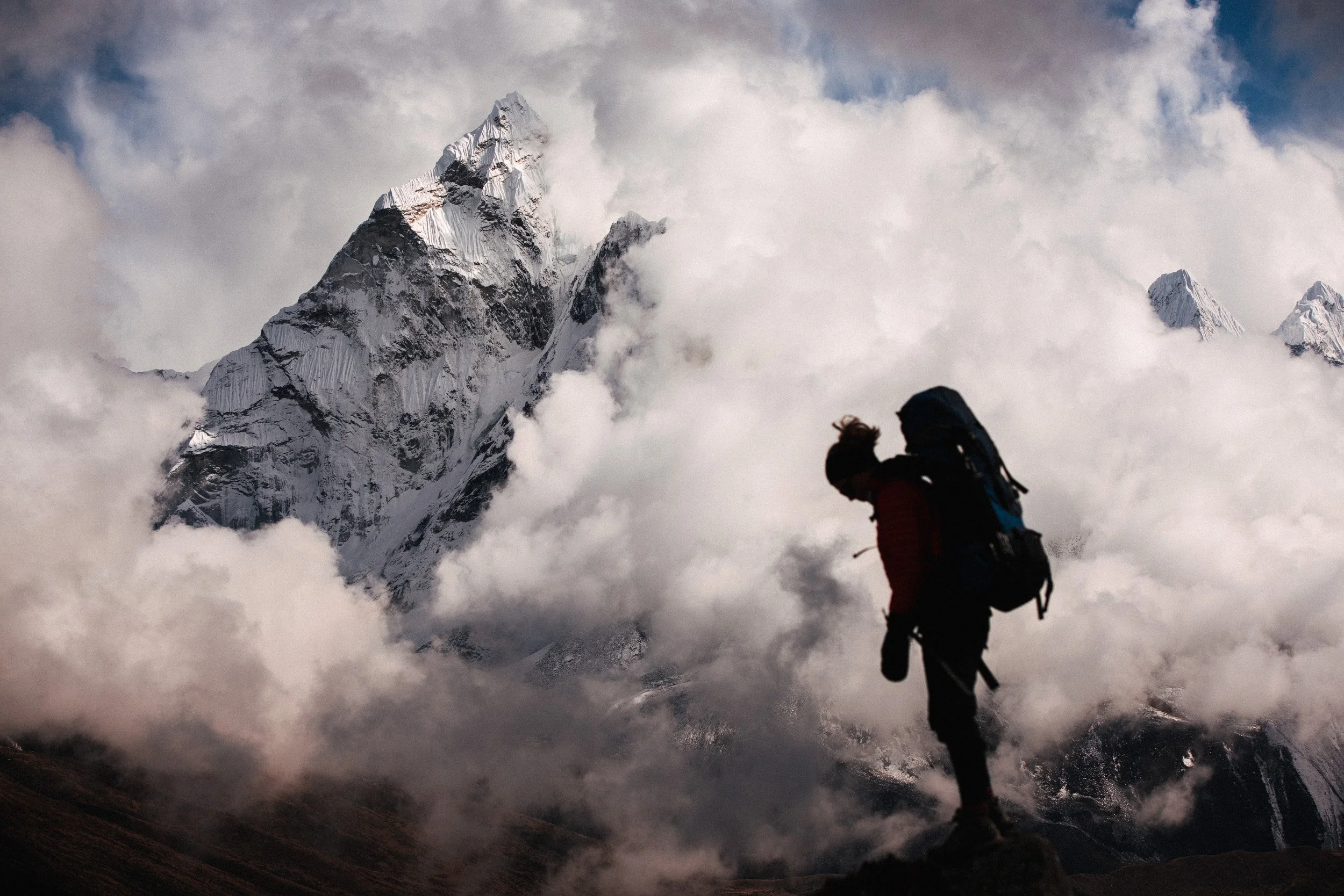 A mountain climber with a backpack standing on rocky terrain, overlooking snow-capped peaks and clouds in a mountainous landscape.